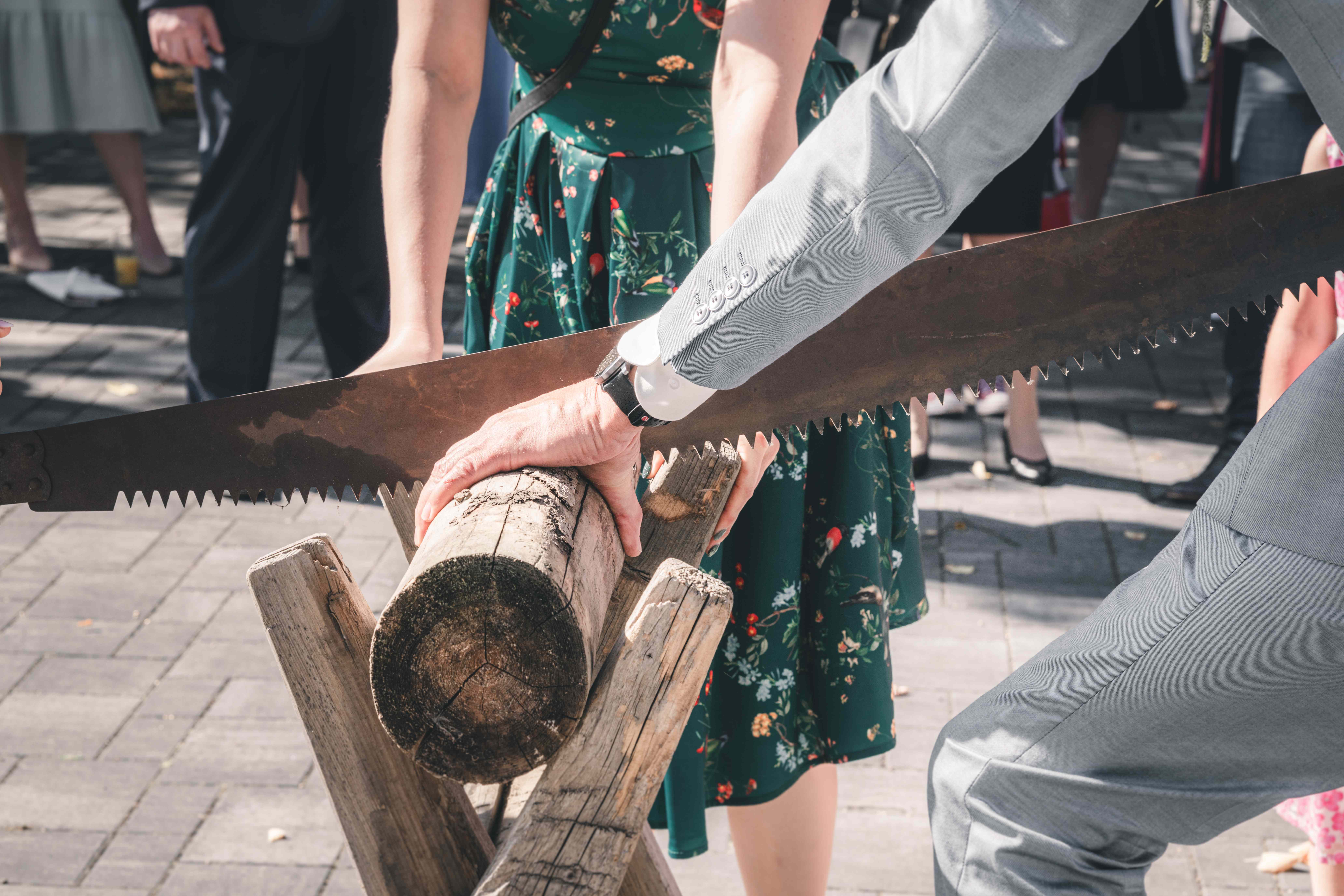 Eine Nahaufnahme des Brautpaares, das gemeinsam die alte Handsäge durch den Holzstamm auf dem Sägebock führt.