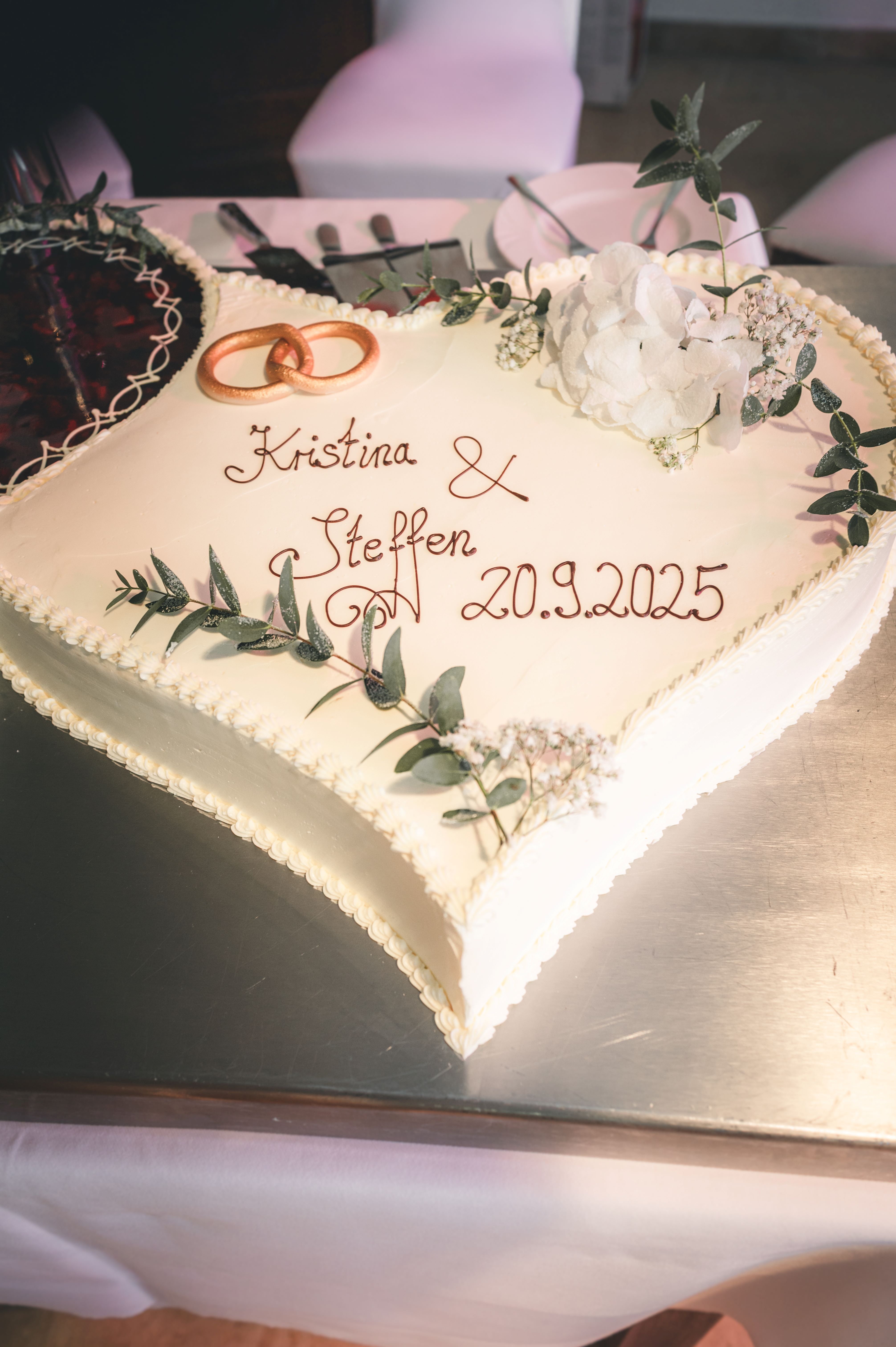 Nahaufnahme der weißen Hochzeitstorte mit der Aufschrift Kristina & Steffen, verziert mit goldenen Ringen und Eukalyptus im Erzgebirge.