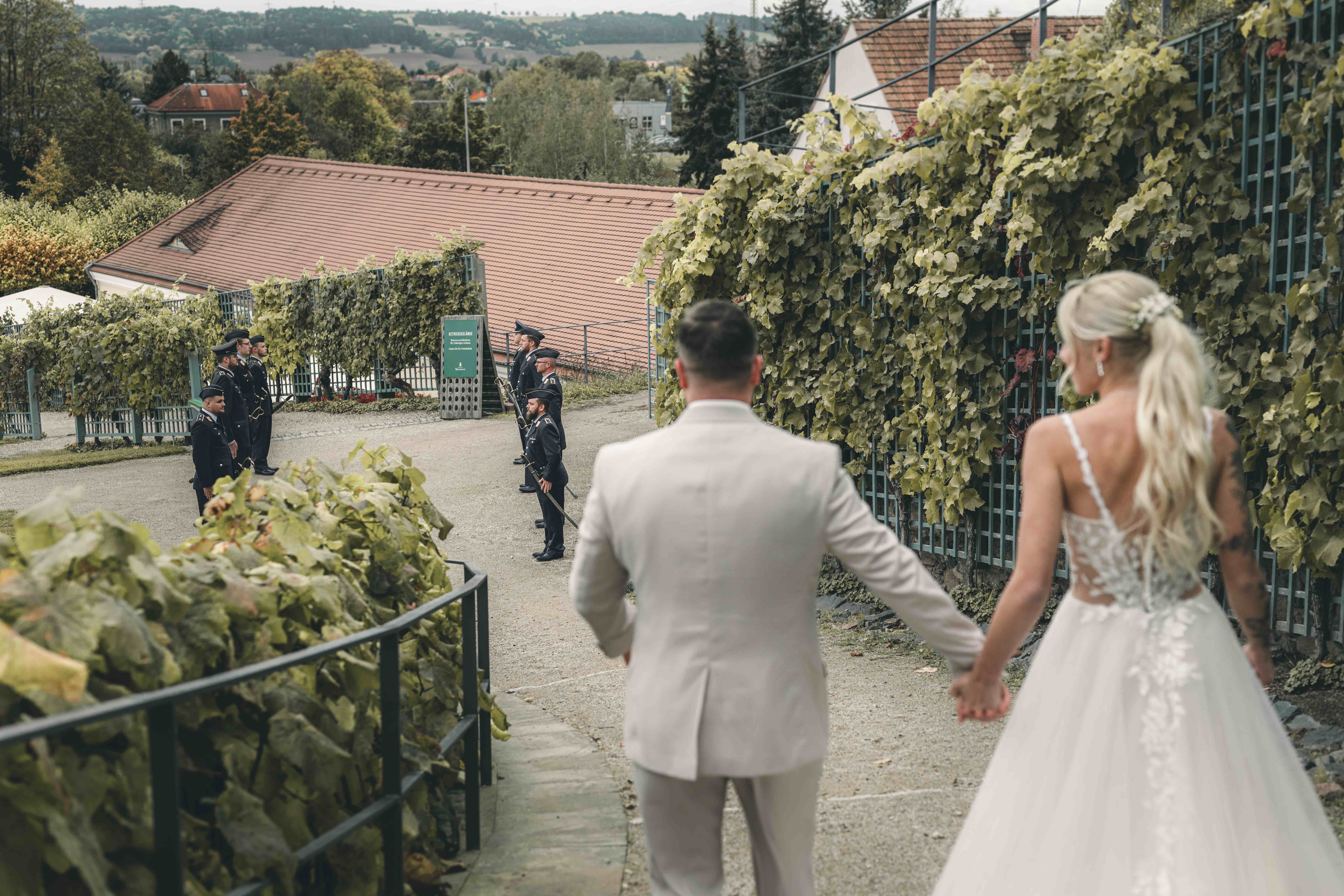 Rückansicht des Brautpaares, das Hand in Hand die gepflasterten Wege von Schloss Wackerbarth hinabläuft, wo in der Ferne uniformierte Soldaten warten.