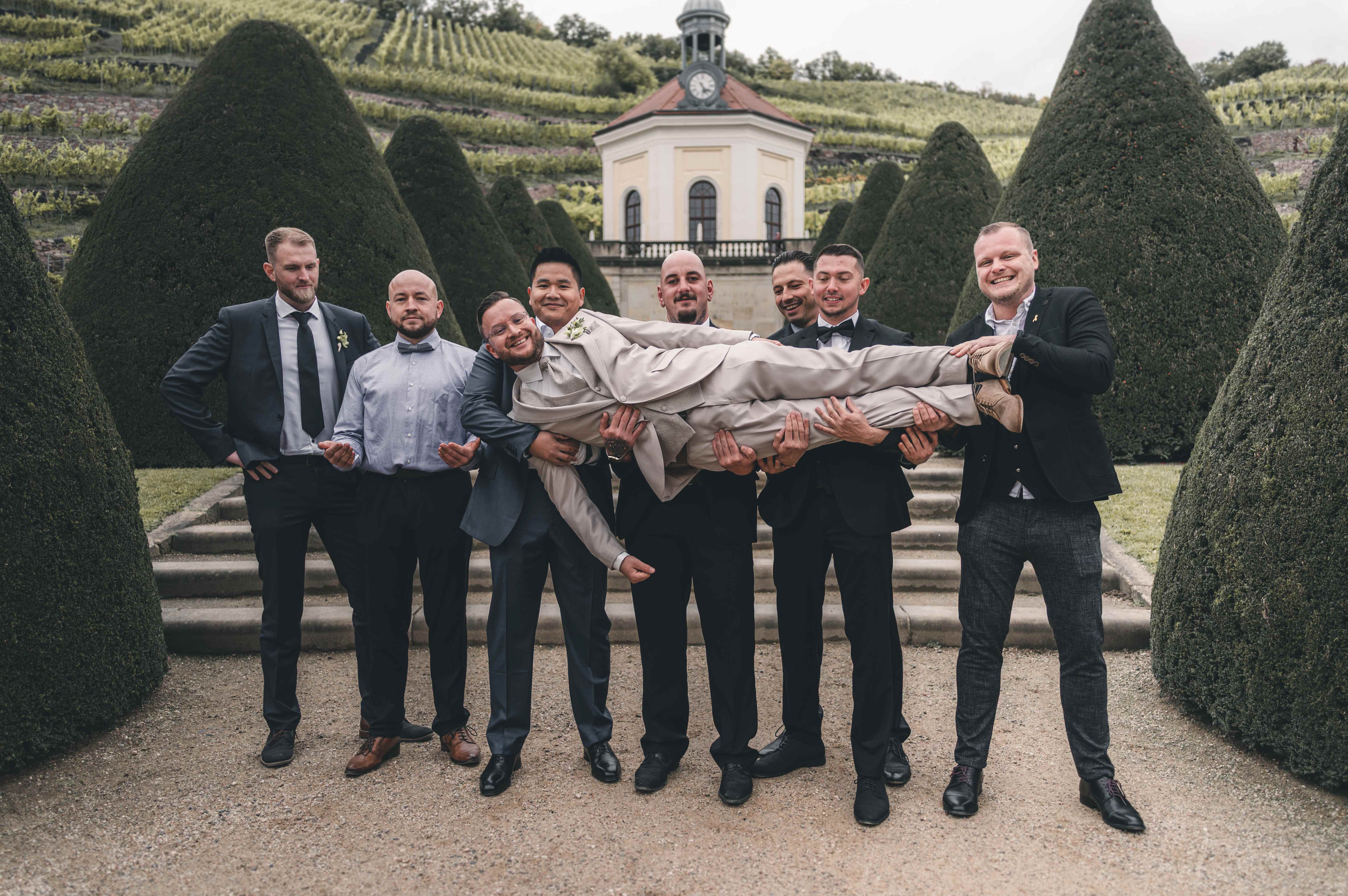 Der Bräutigam wird von seinen Trauzeugen und Freunden im Barockgarten von Schloss Wackerbarth scherzhaft auf Händen getragen, im Hintergrund steht das Belvedere.