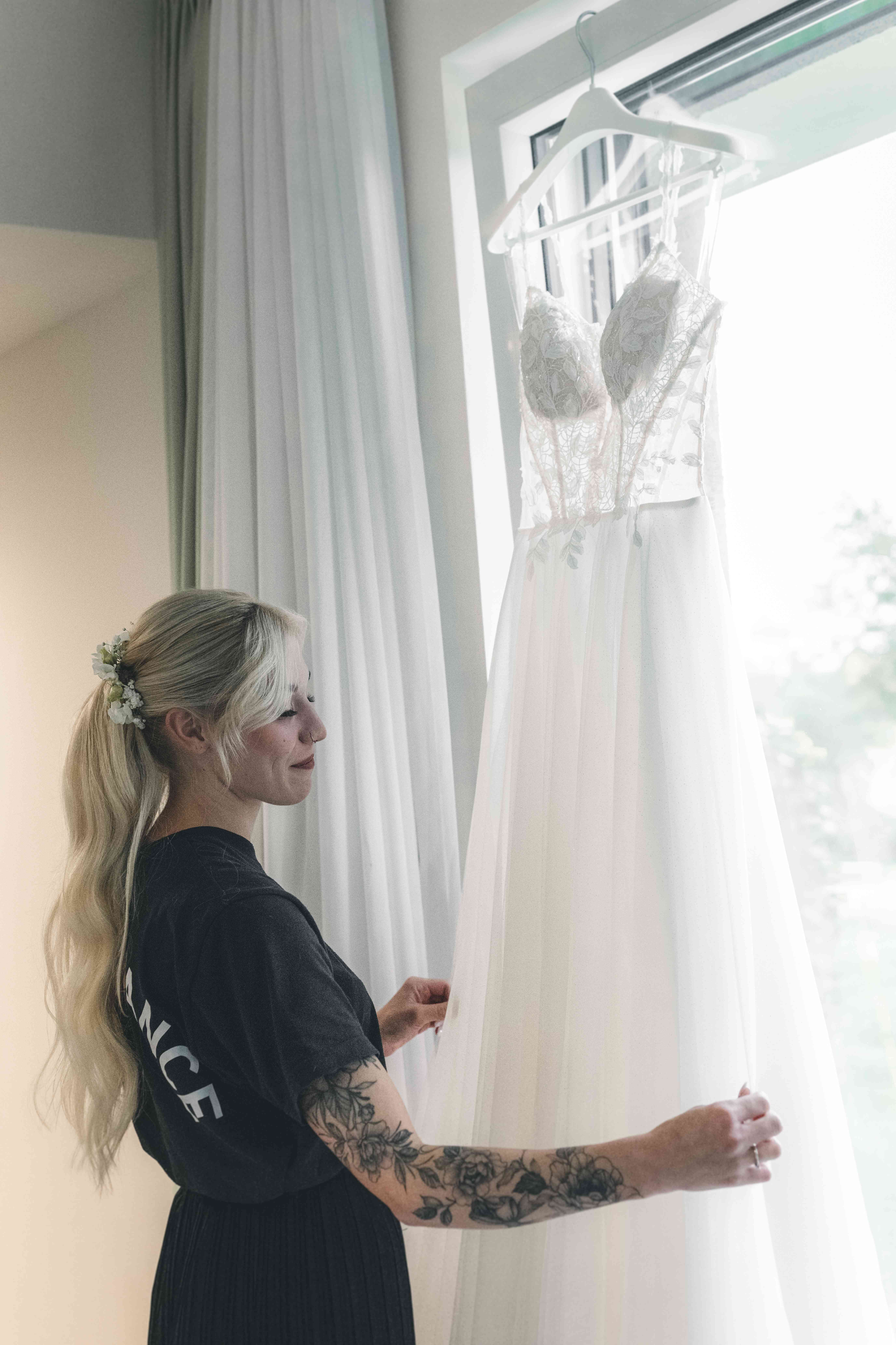 Ein berührendes Porträt der Braut im Profil, die sanft lächelnd den Tüll-Stoff ihres am Fenster hängenden Hochzeitskleids berührt.