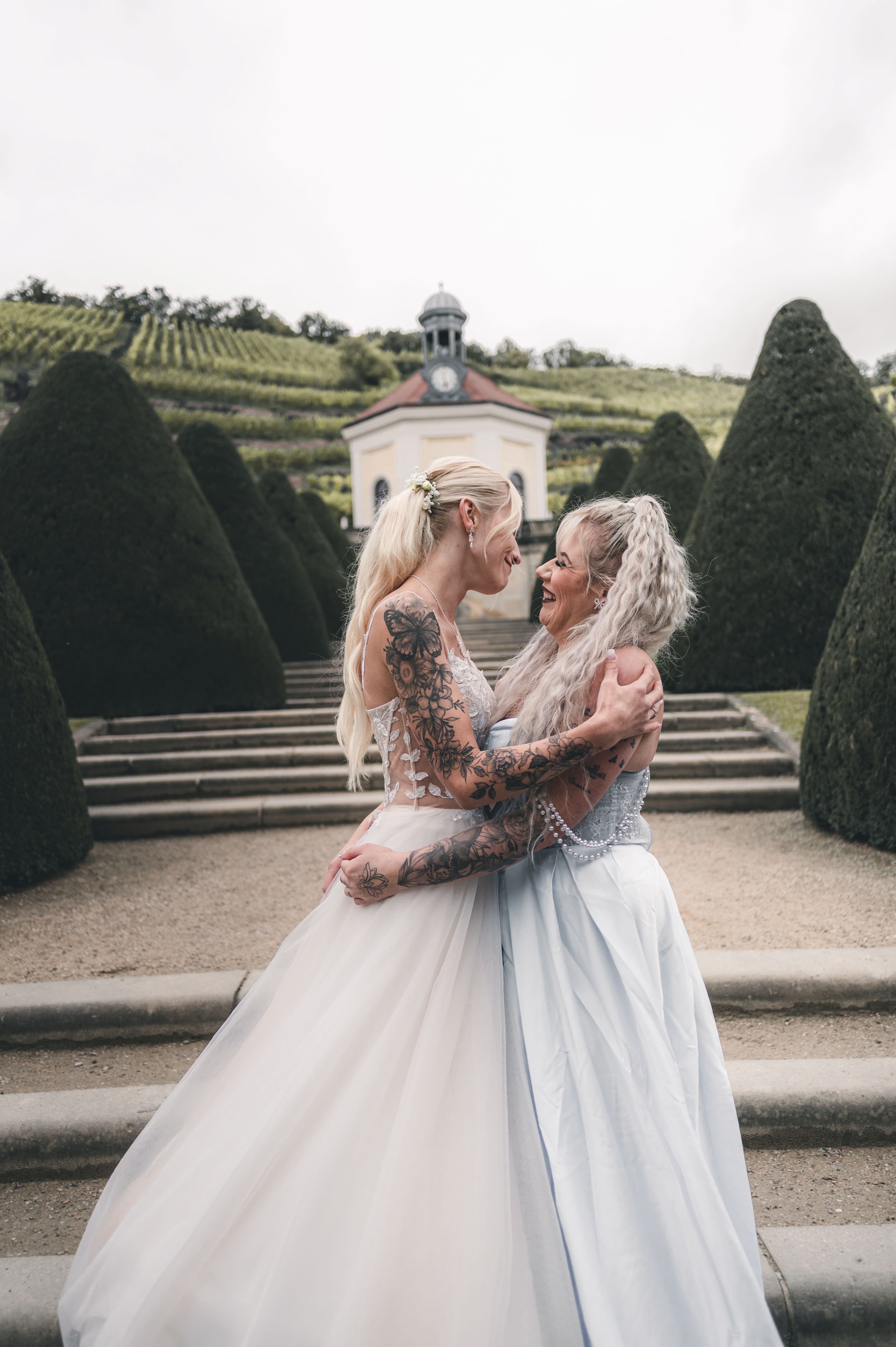 Die Braut liegt einer engen Vertrauten oder ihrer Schwester im Schlossgarten glücklich in den Armen, im Hintergrund sind die berühmten Weinhänge zu sehen.