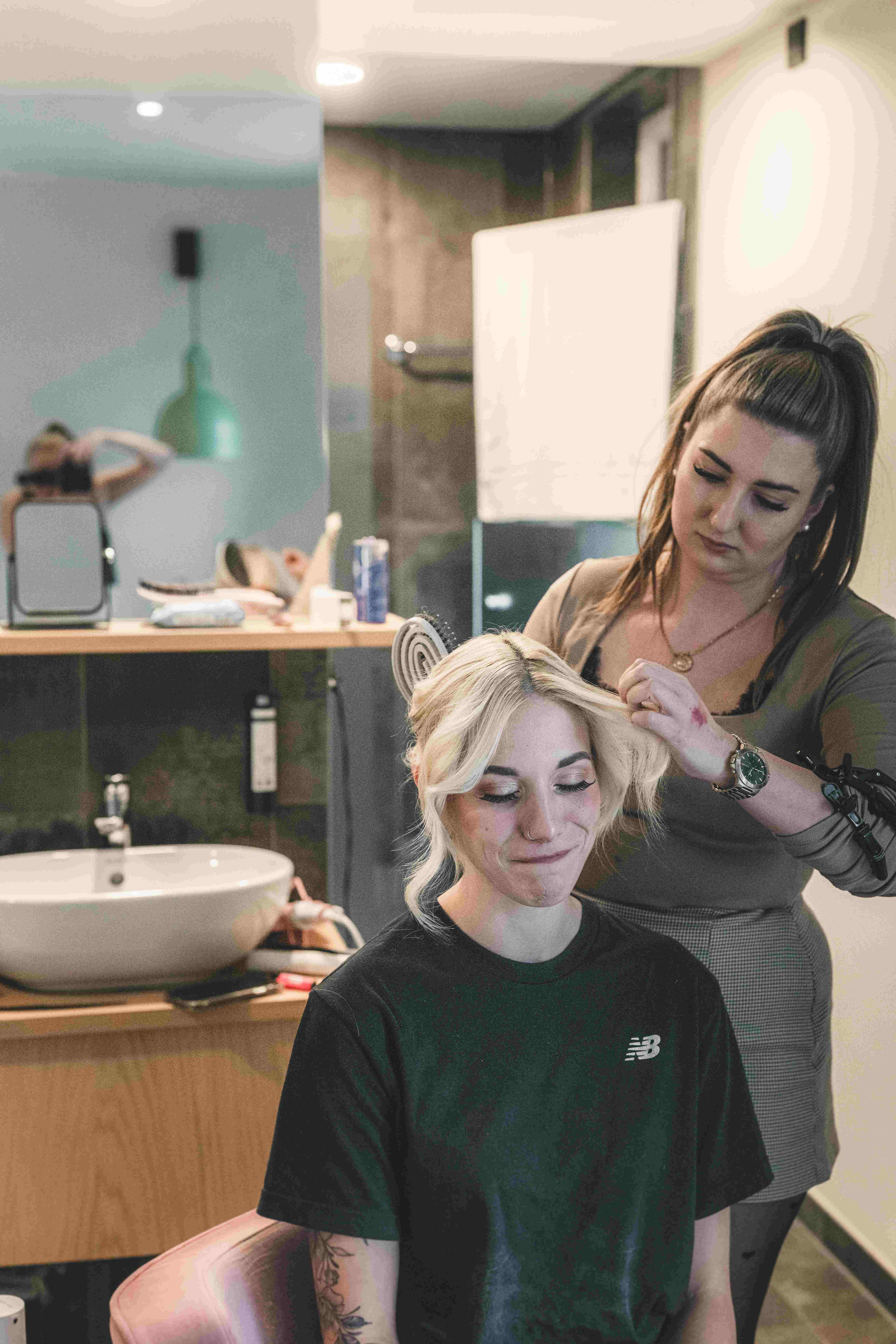 Eine Braut lächelt entspannt im hellen Zimmer, während die Hairstylistin ihre blonden Haare zu eleganten Locken für die Trauung formt.