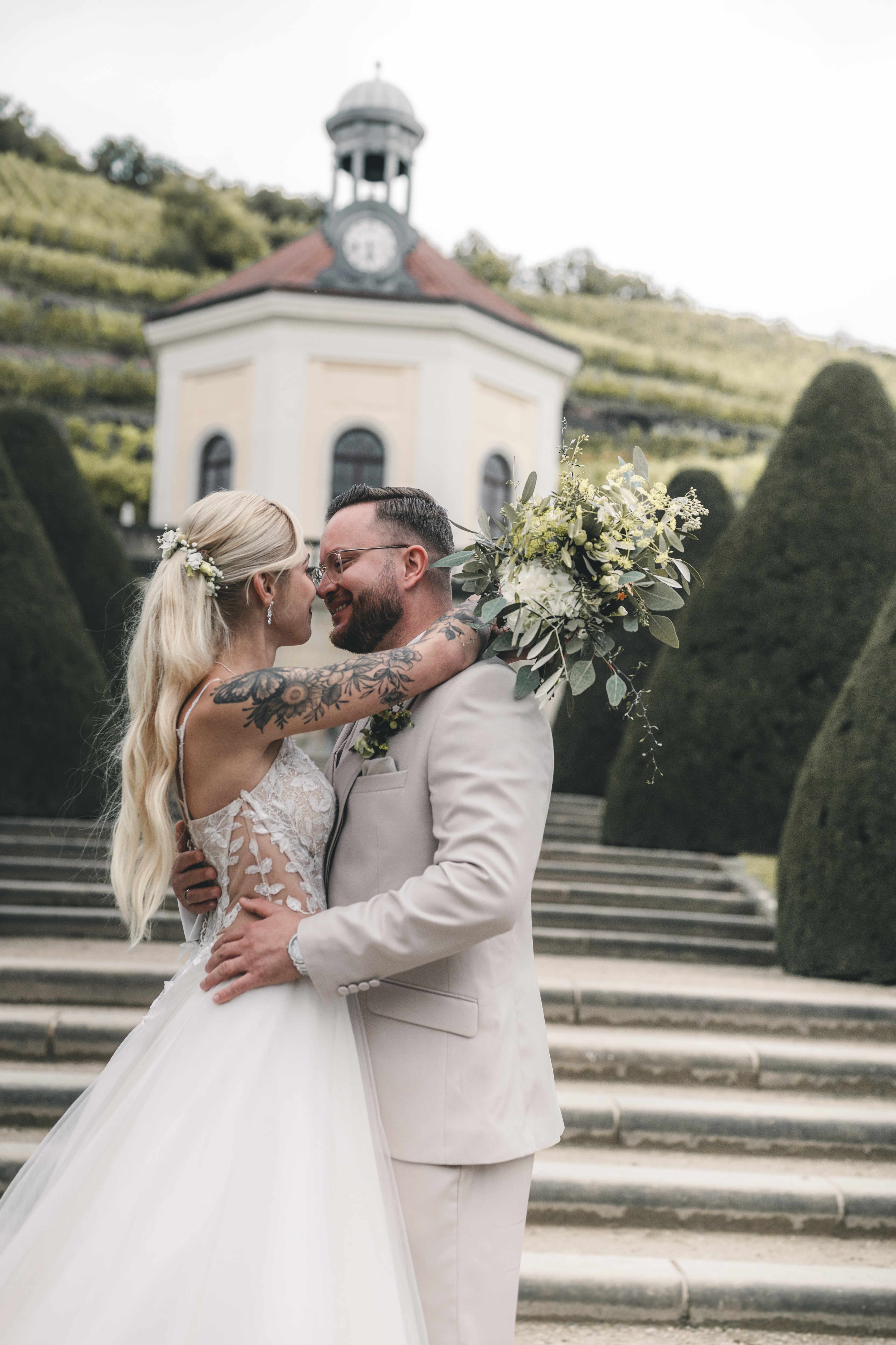 Braut und Bräutigam stehen eng umschlungen auf den Steinstufen vor dem Belvedere-Pavillon auf Schloss Wackerbarth und halten den Brautstrauß.
