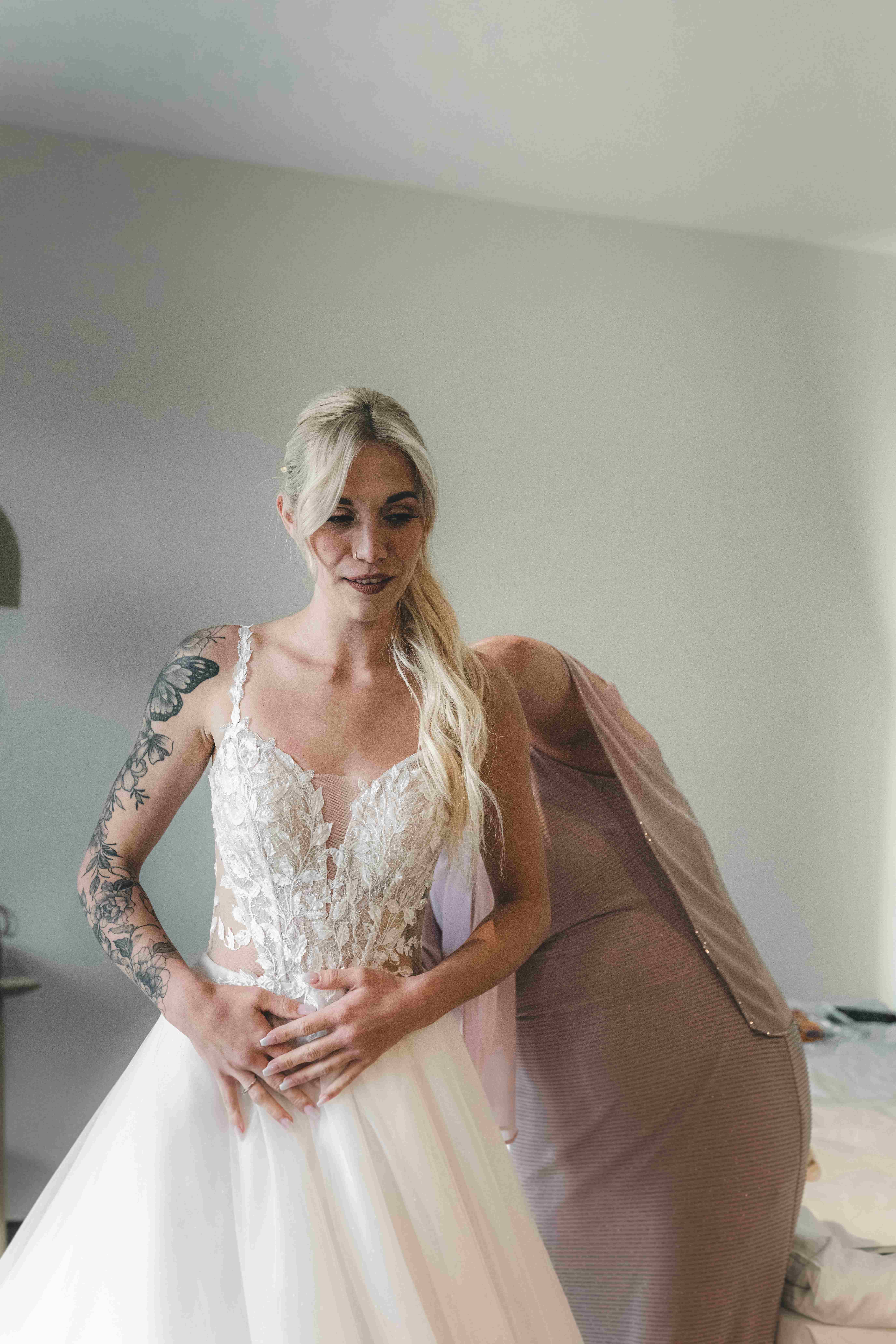 Porträt der fertig gestylten Braut im Spitzenkleid mit floralen Trägern, die Hände sanft auf den Bauch gelegt, mit sichtbaren Arm-Tattoos.