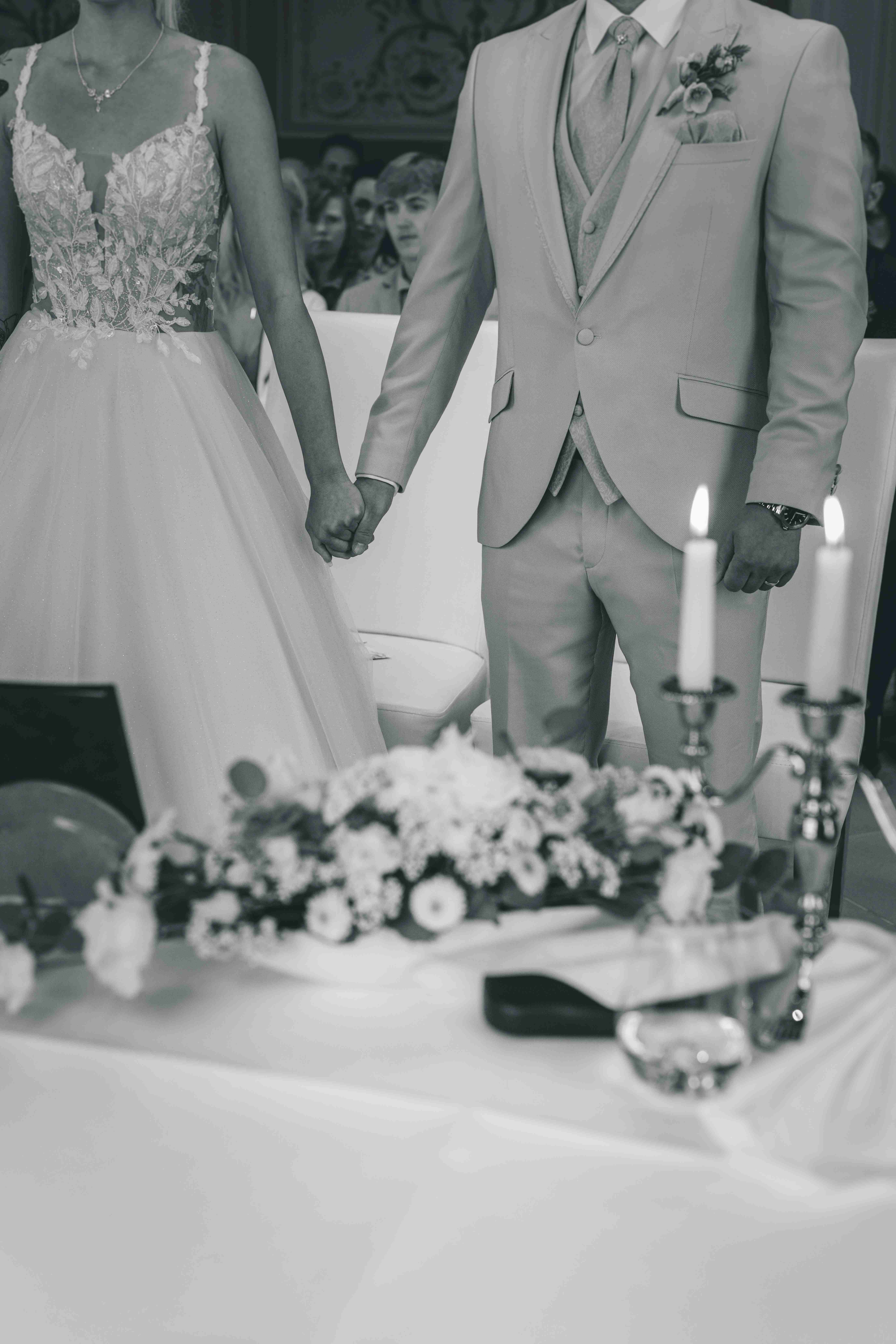 Schwarz-Weiß-Detailaufnahme des Brautpaares, das sich am Hochzeitstisch im Standesamt eng an den Händen hält und die neuen Eheringe zeigt.