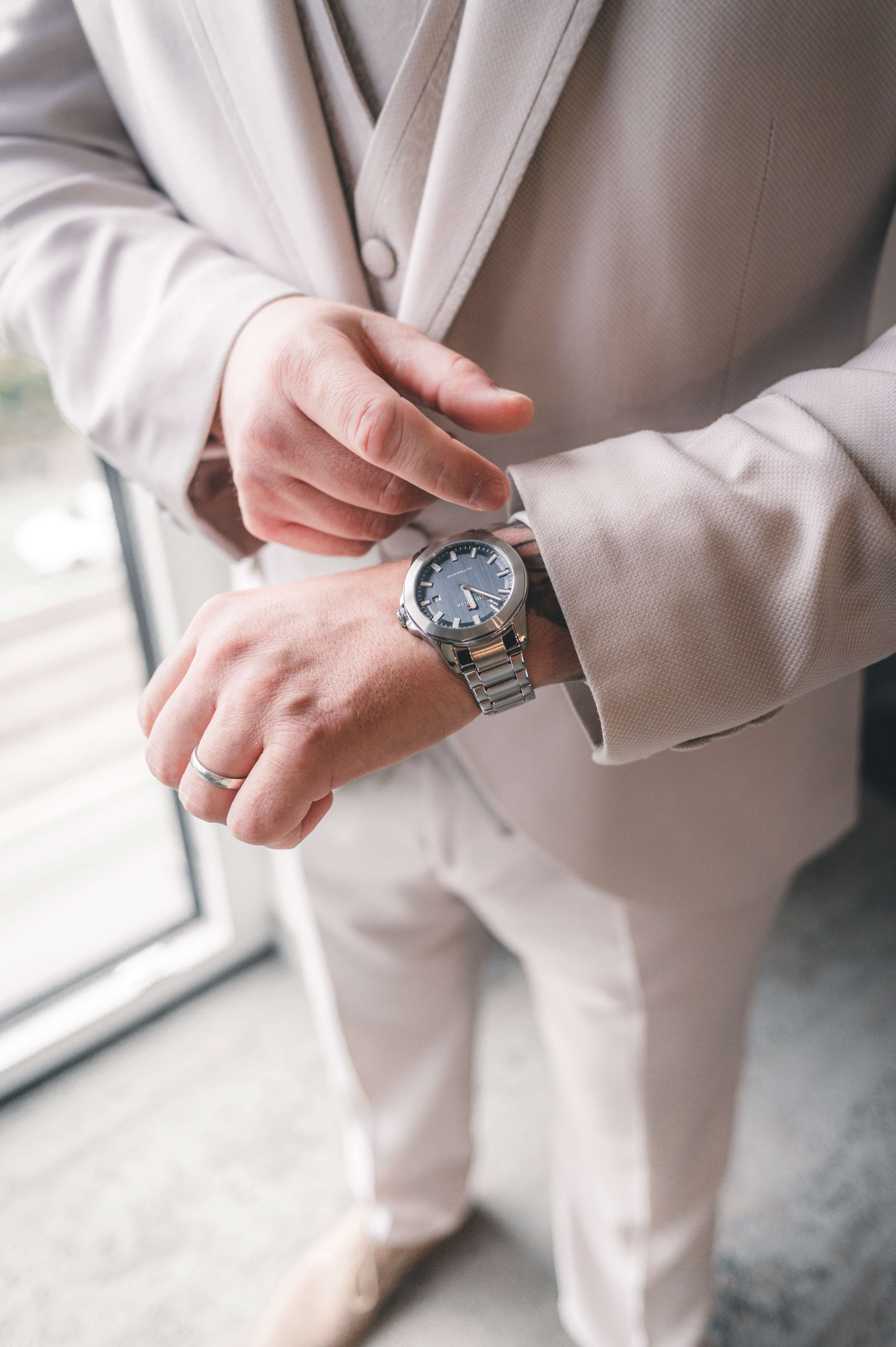 Nahaufnahme eines Bräutigams, der im hellen Anzug auf seine silberne Armbanduhr blickt, während sein Ehering am Finger funkelt.