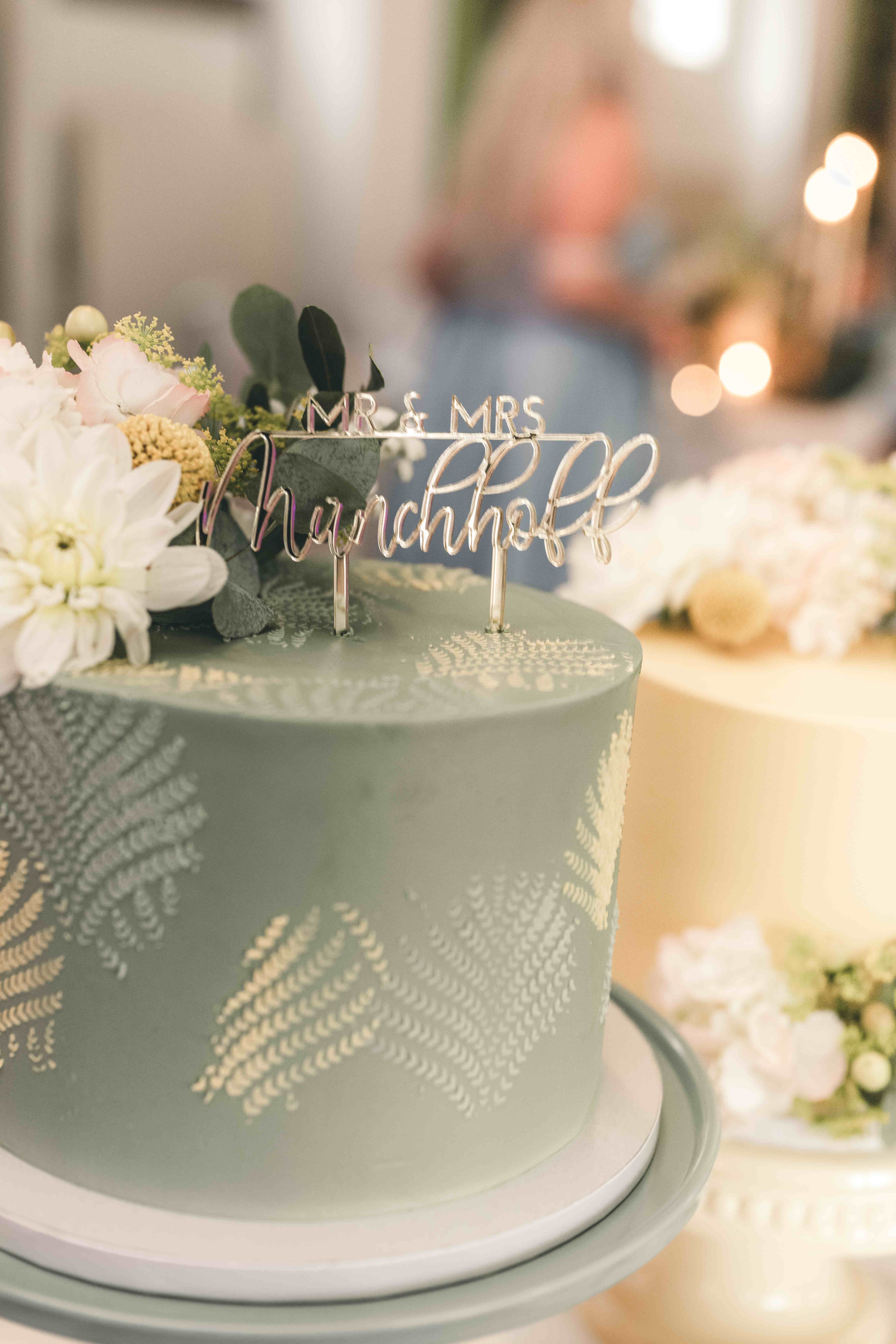 Detailaufnahme einer salbeigrünen Hochzeitstorte mit filigranem Farn-Muster und einem eleganten silbernen Cake Topper mit der Aufschrift „Mr. & Mrs. Munchhoff“.