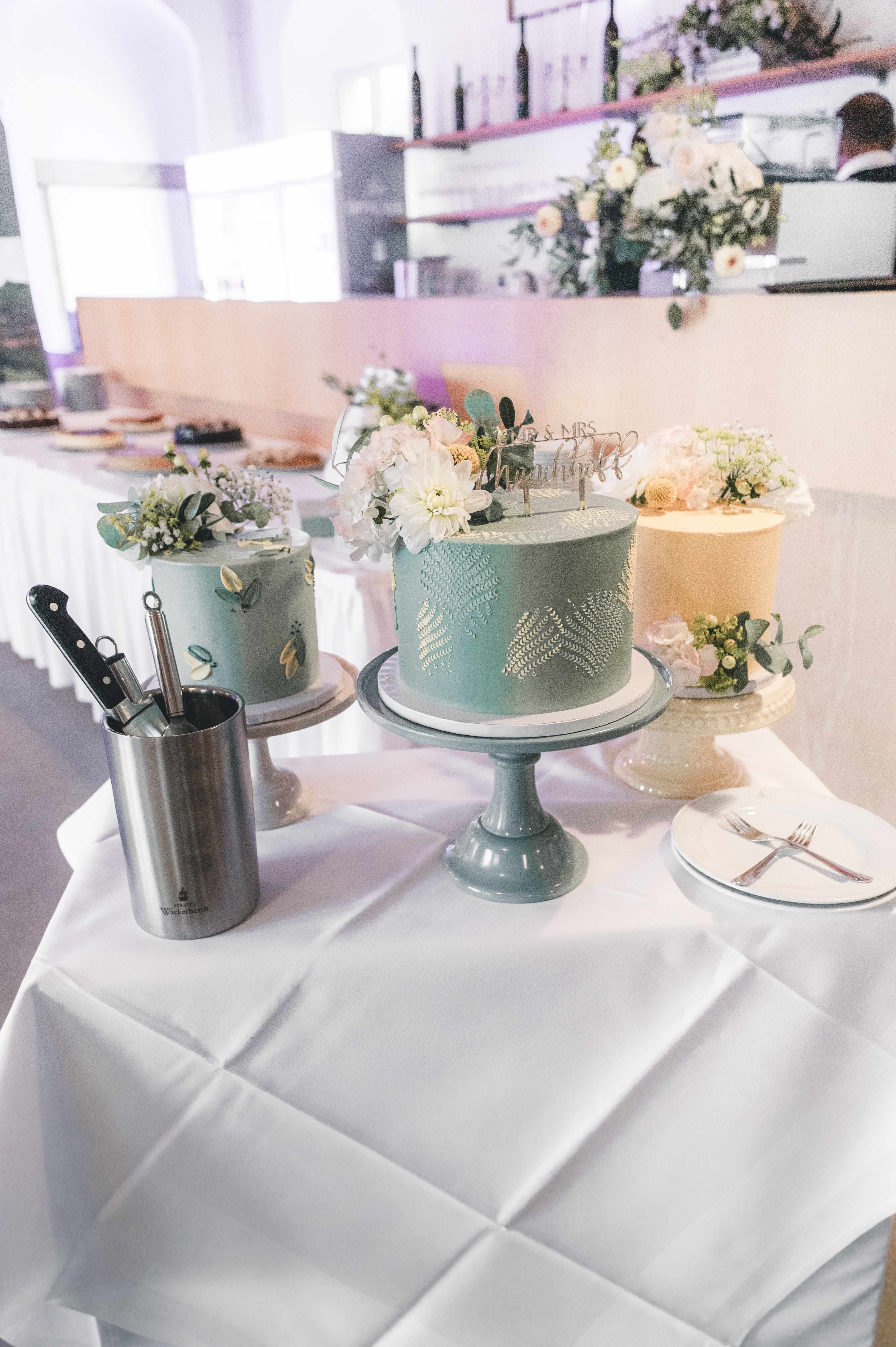 Drei einzeln arrangierte Hochzeitstorten in Salbeigrün und Creme, dekoriert mit echten weißen Blumen, stehen bereit für den Tortenanschnitt im Mörtelwerk-Stil auf Schloss Wackerbarth.