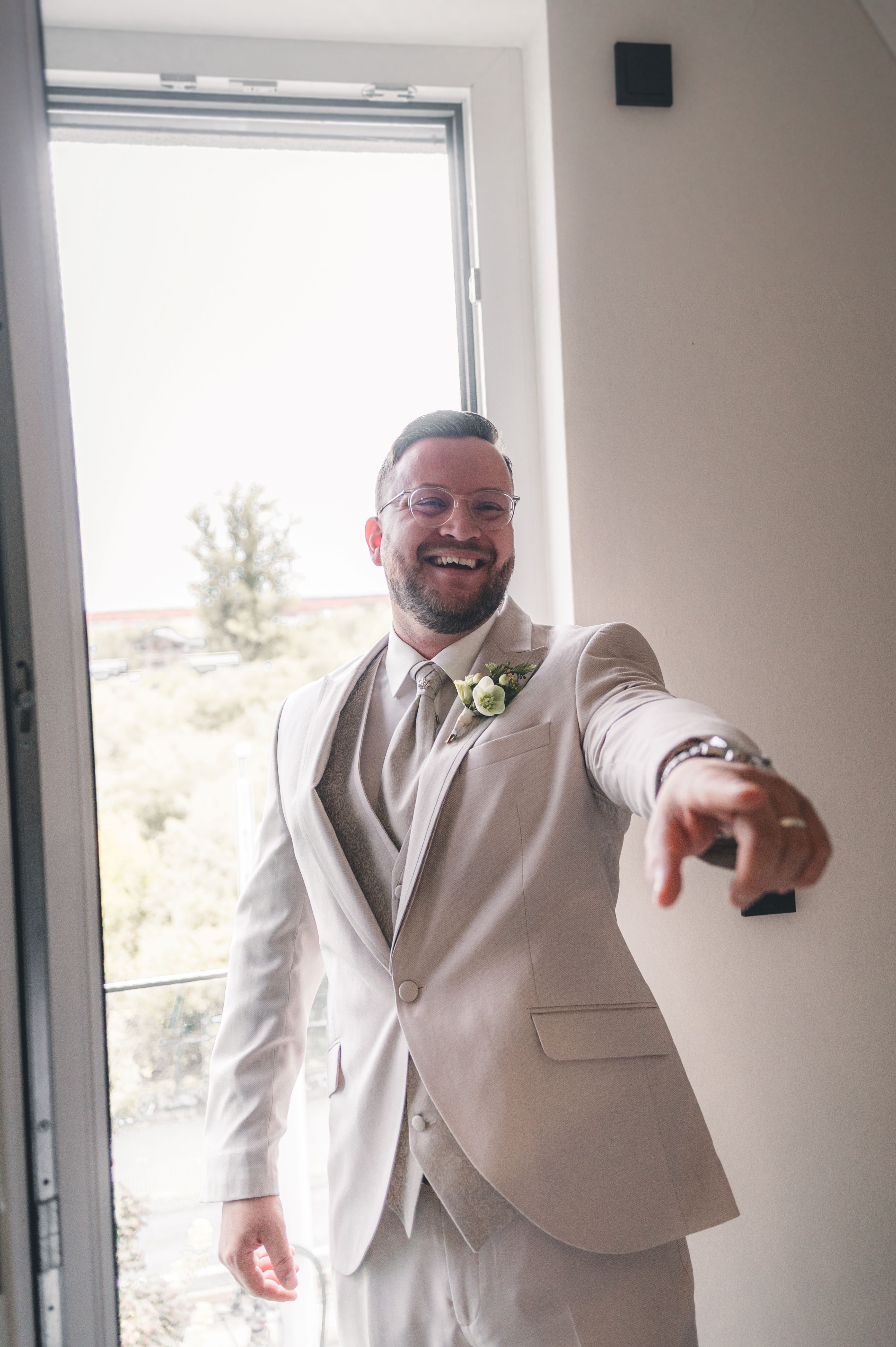 Ein strahlender Bräutigam mit Brille im eleganten, hellbeigen Hochzeitsanzug mit Weste und Krawatte zeigt gut gelaunt in die Kamera.