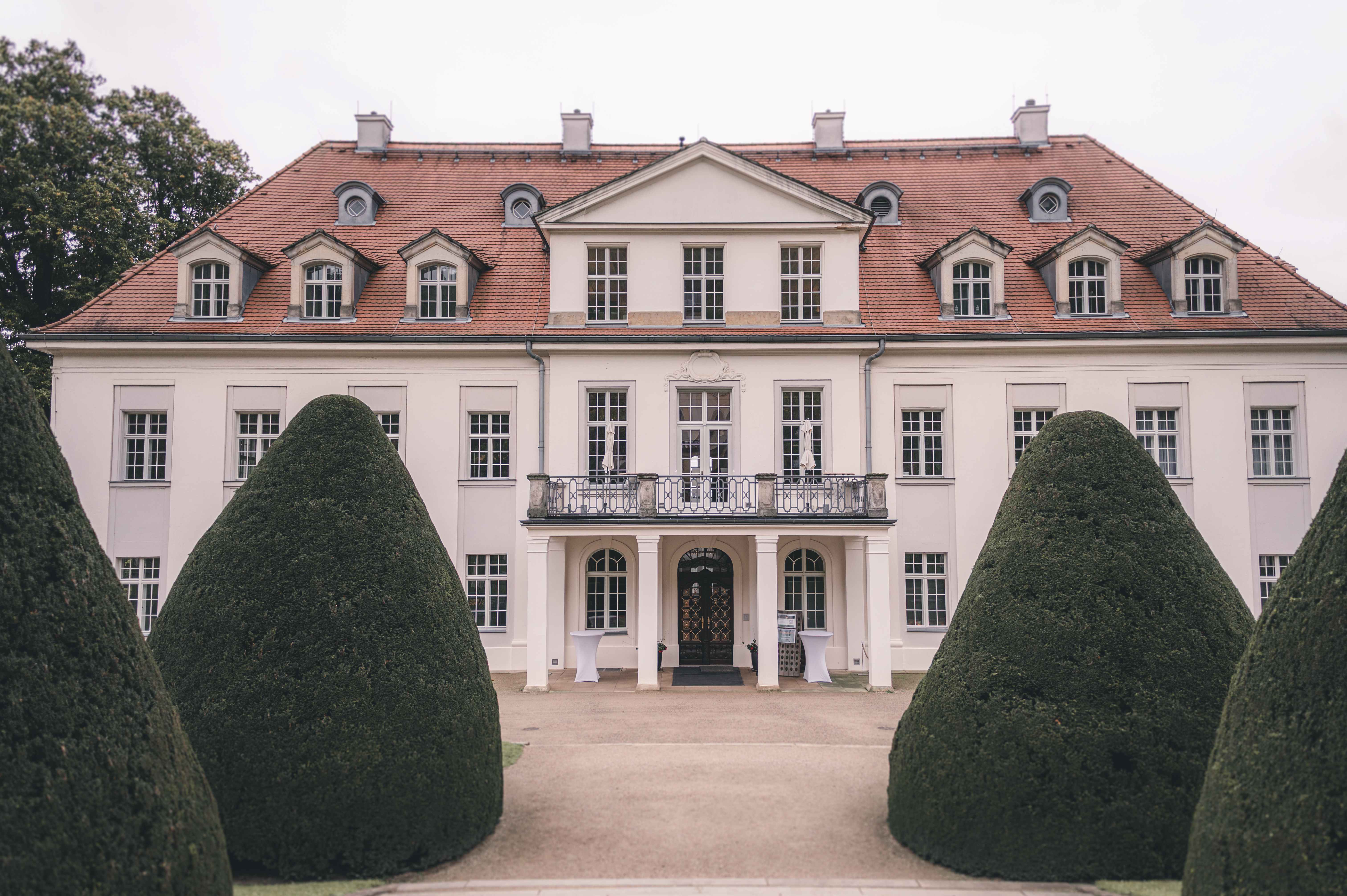 Symmetrische Außenaufnahme der barocken Schloss-Fassade von Schloss Wackerbarth, gerahmt von kegelförmig geschnittenen Hecken im Schlossgarten.