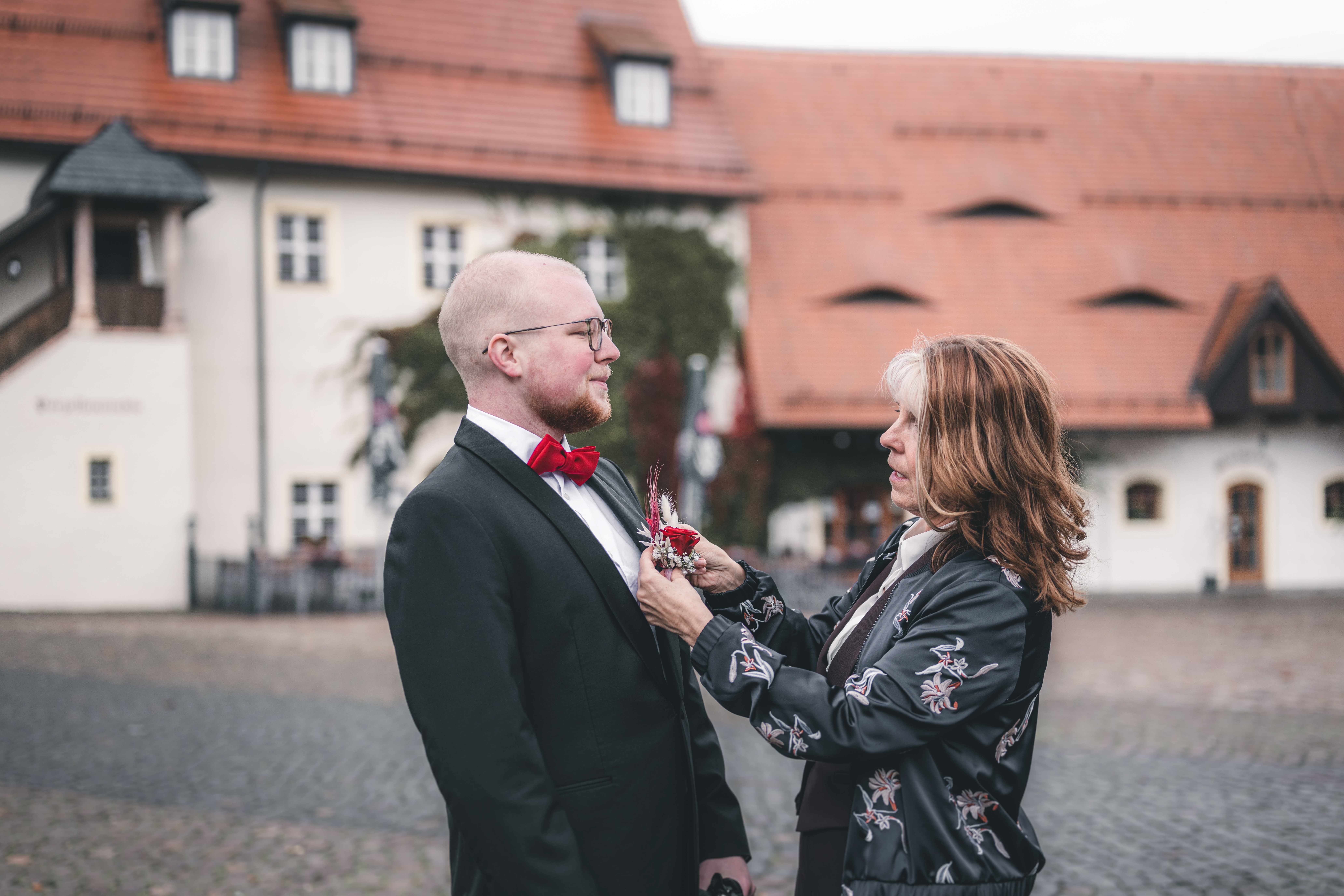 Eine Gästin befestigt dem Bräutigam im schwarzen Anzug mit roter Fliege die Ansteckrose vor der Kulisse des Schlossgasthofes Klaffenbach.
