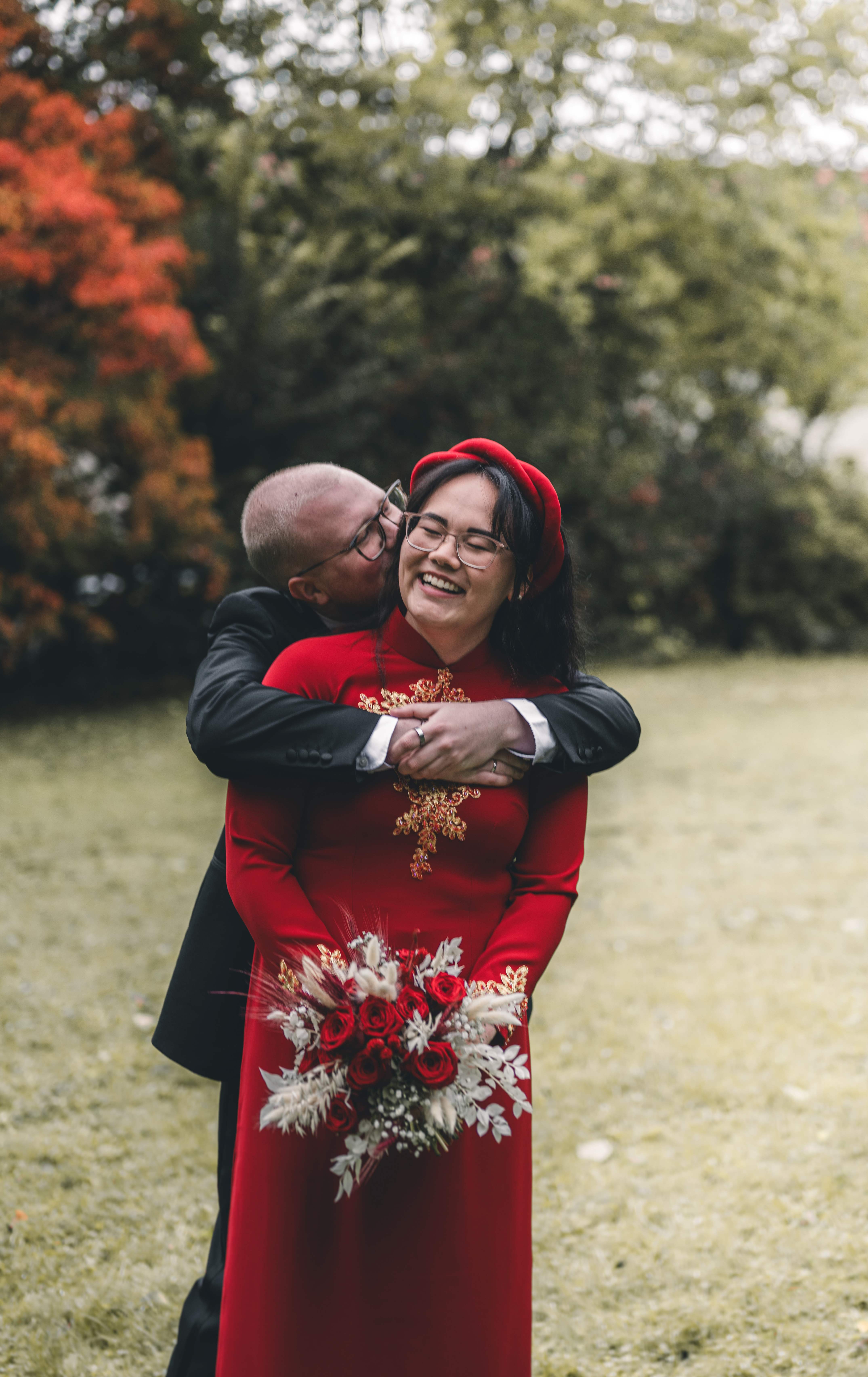 Der Bräutigam umarmt seine Braut von hinten, während sie im roten Ao Dai glücklich lächelnd ihren roten Rosenstrauß hält.