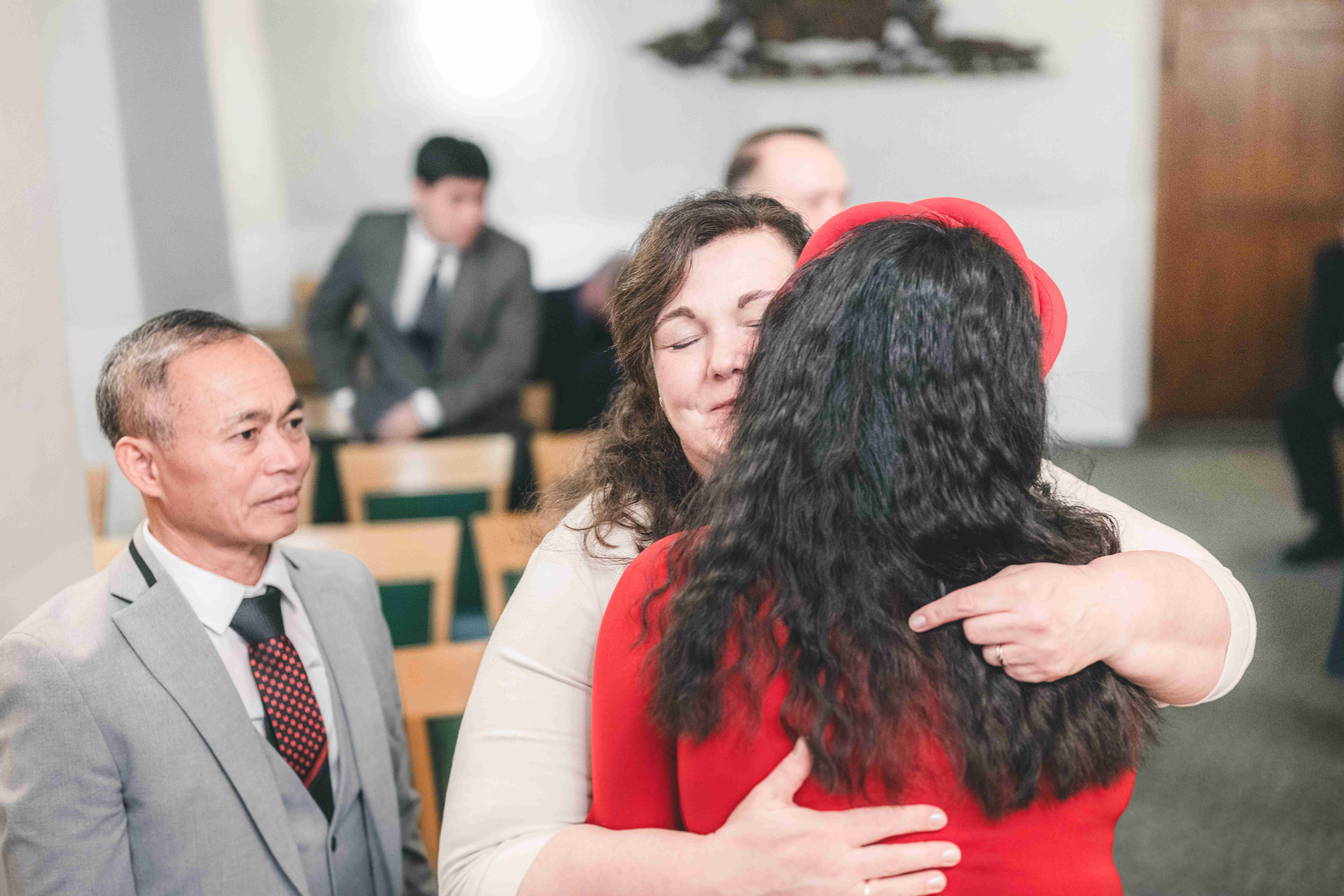 Eine Gästin schließt die Braut im roten Kleid nach der Trauung im herrschaftlichen Saal für eine feste Umarmung gerührt in die Arme.