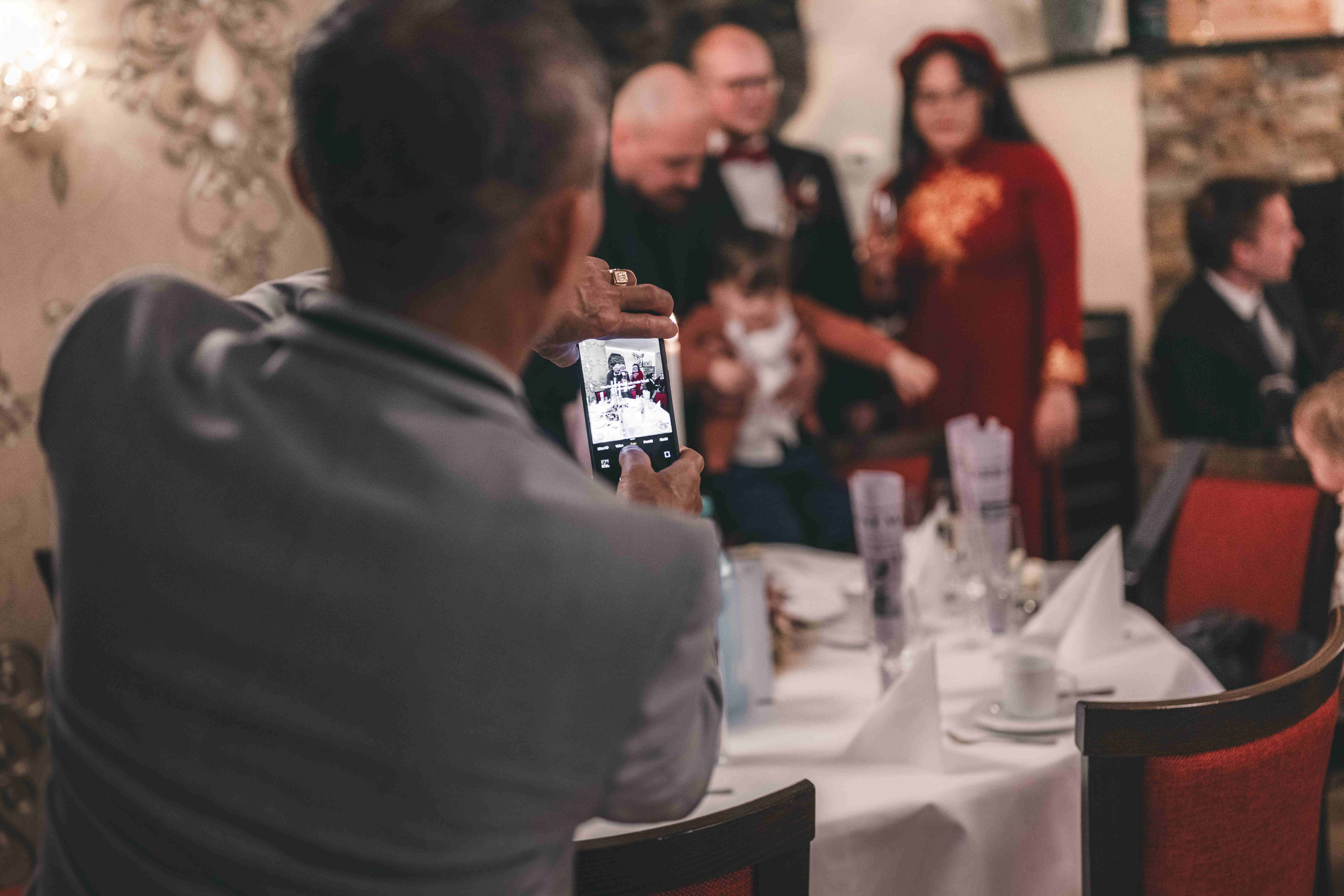 Die Perspektive über die Schulter eines Hochzeitsgastes, der im festlichen Saal mit seinem Smartphone ein Foto von der Familie macht.