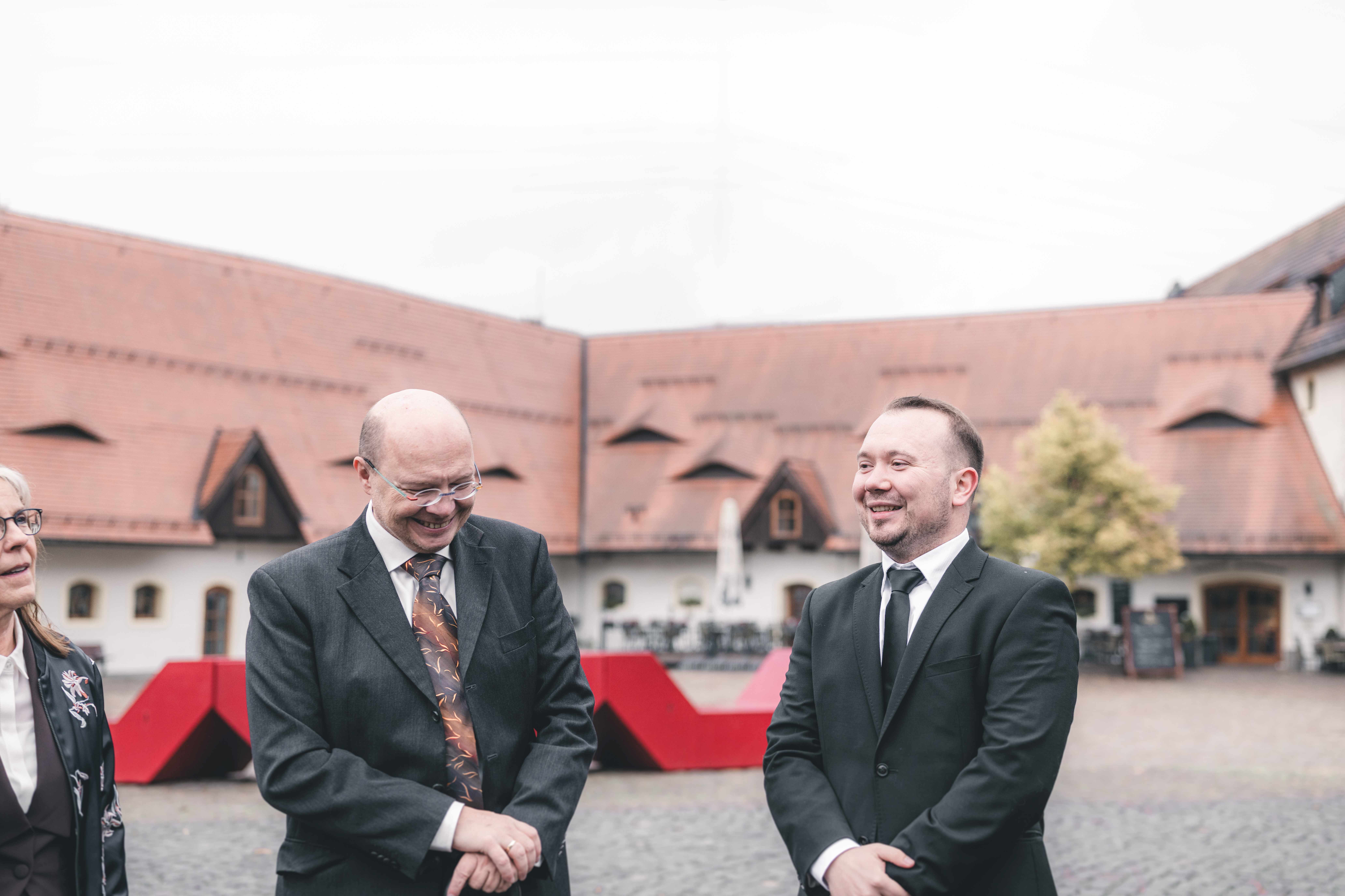 Zwei elegant gekleidete Hochzeitsgäfte unterhalten sich gut gelaunt und lachend im großen Pflasterinnenhof des Wasserschlosses Klaffenbach.