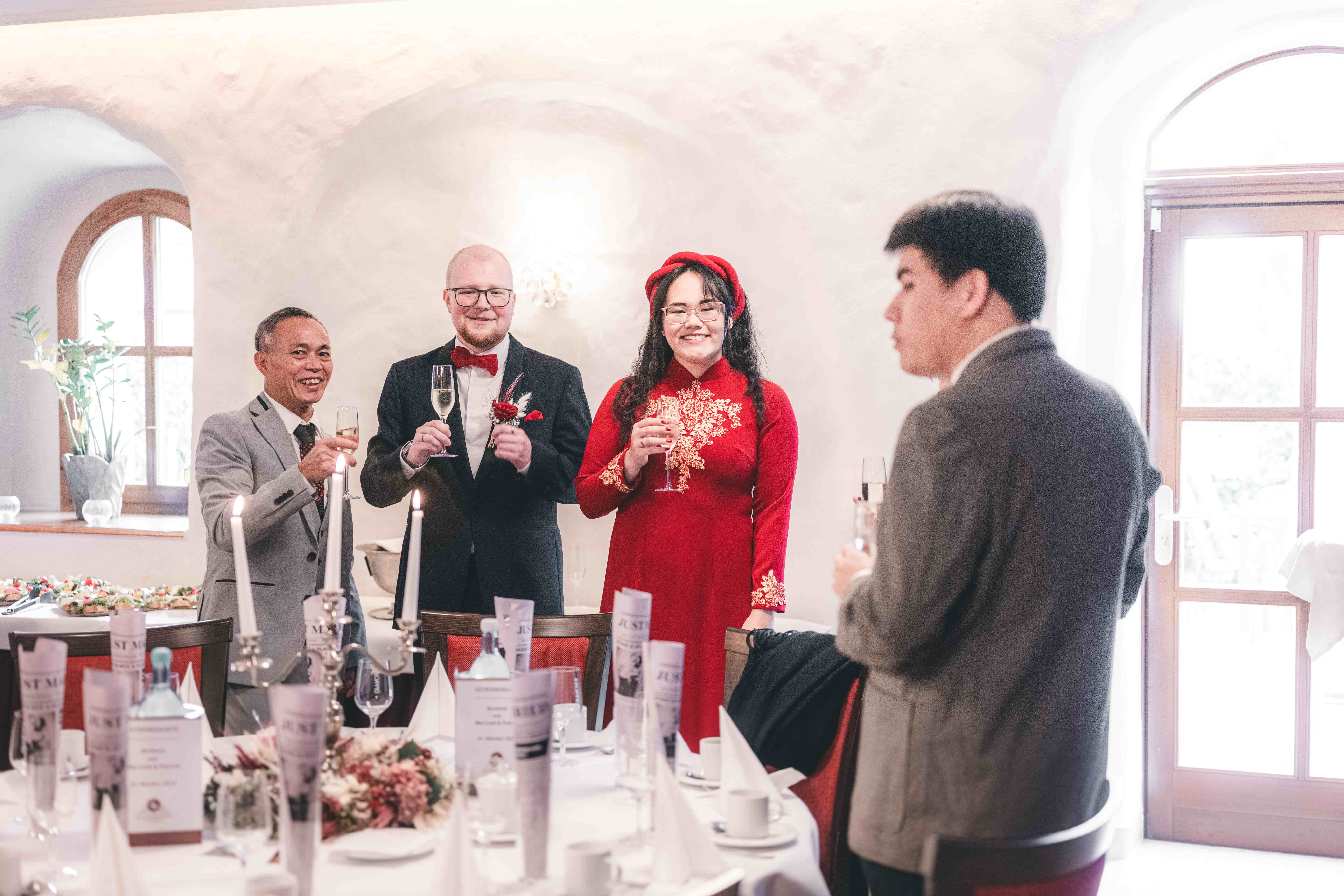 Das Brautpaar in Hochzeitskleidung steht lächelnd mit Gästen am Tisch und erhebt die Sektgläser zum gemeinsamen Toast im Saal.