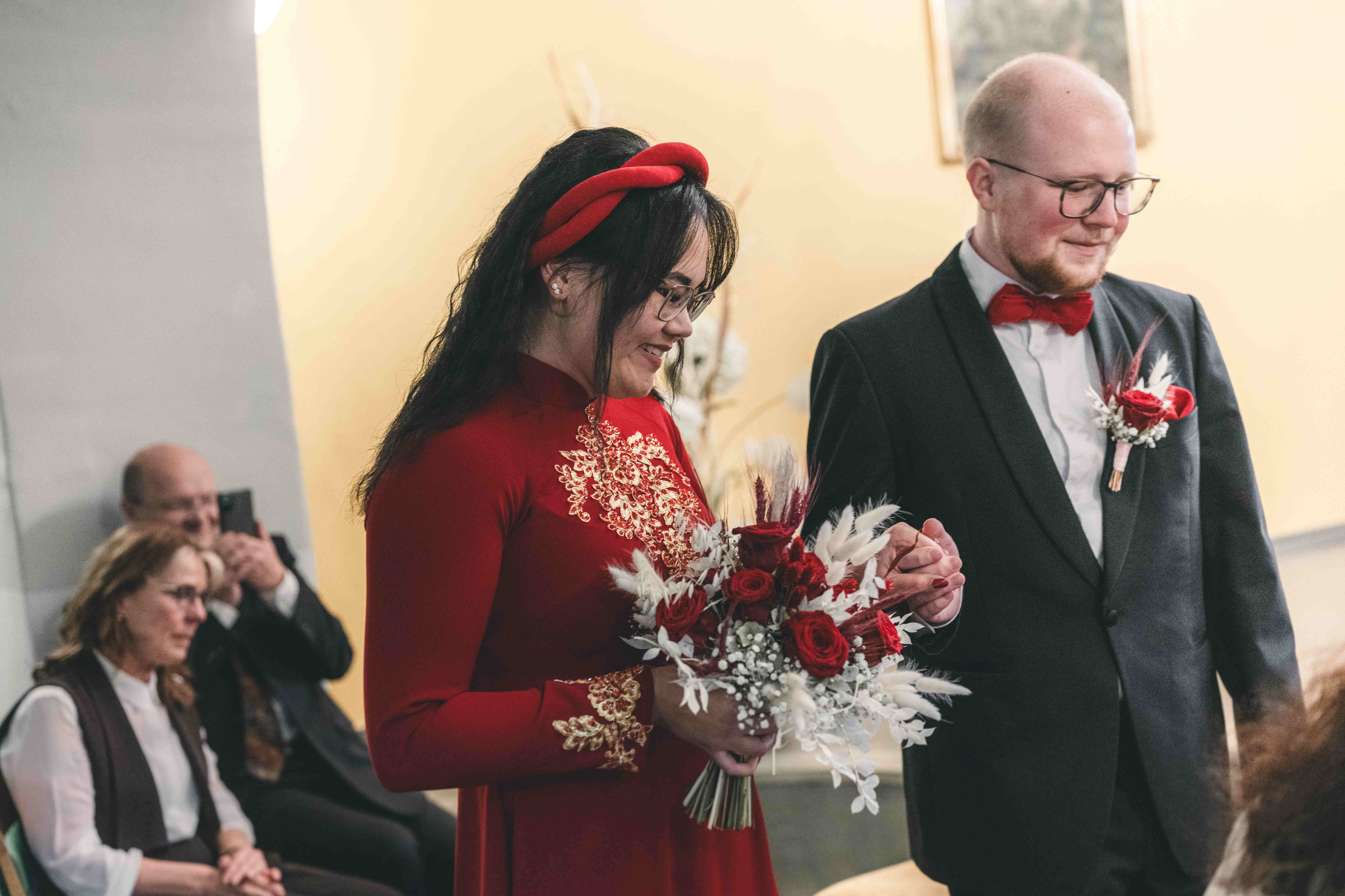 Das Brautpaar sitzt Hand in Hand im Trausaal von Schloss Klaffenbach, die Braut im bestickten roten Kleid lächelt gerührt auf ihren Strauß.