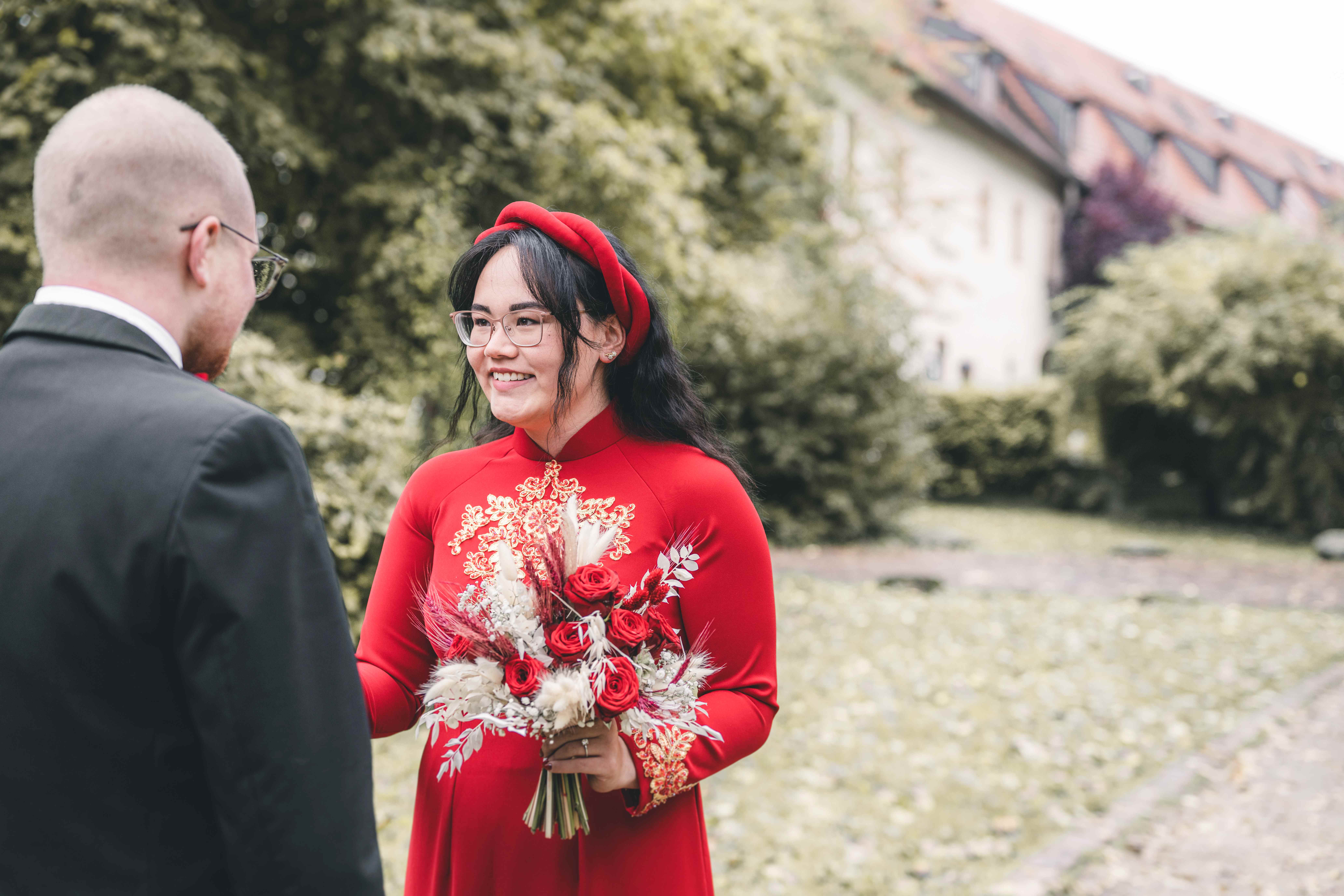 Nahaufnahme der glücklich lächelnden Braut im roten vietnamesischen Ao Dai mit goldenen Stickereien und ihrem rot-weißen Blumenstrauß bei Chemnitz.