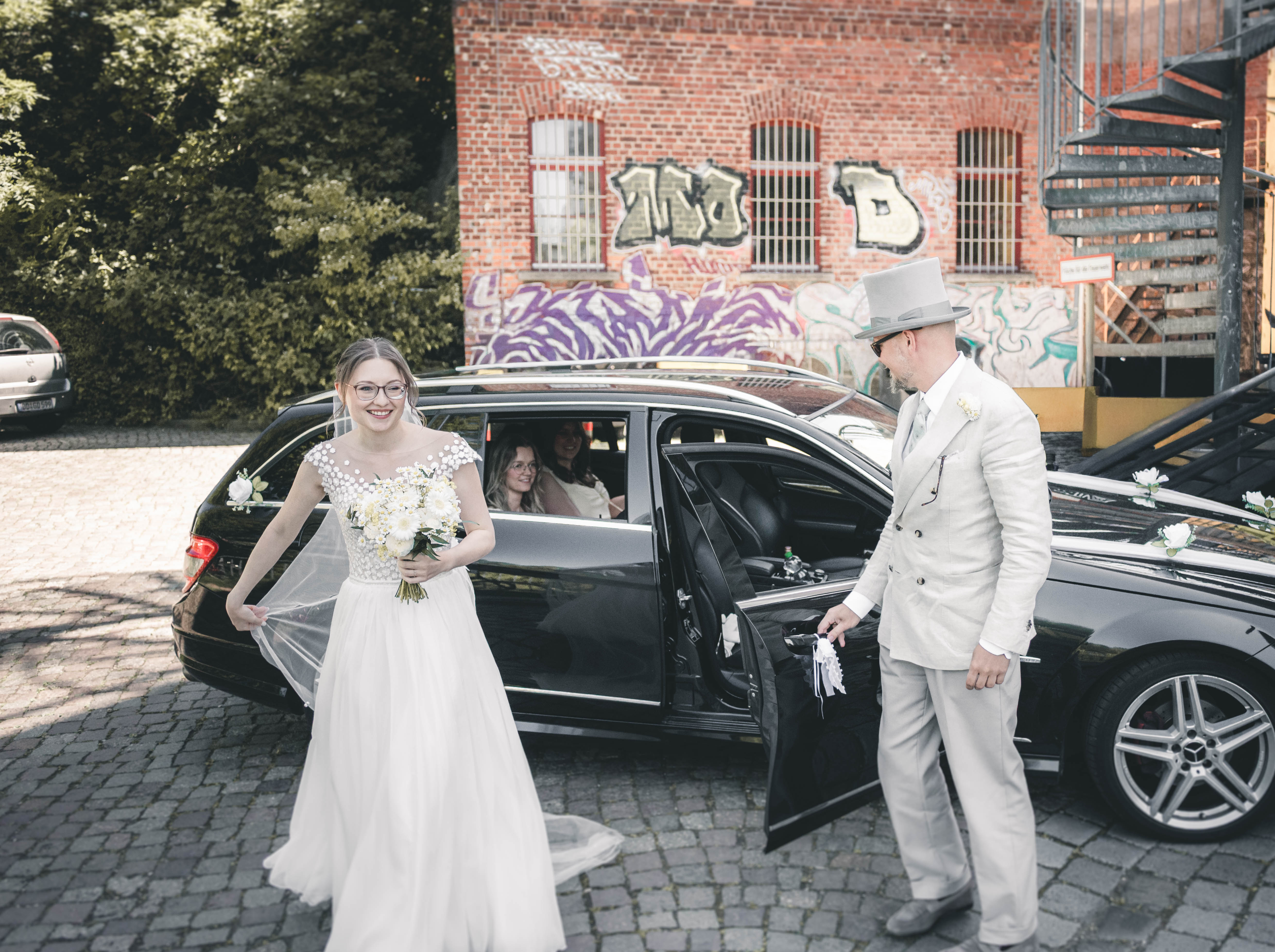 Die strahlende Braut Maja steigt vor der urbanen Graffiti-Backsteinfassade des Mörtelwerks Leipzig aus dem Hochzeitsauto, während ihr ein elegant gekleideter Herr im Frack die Tür aufhält.