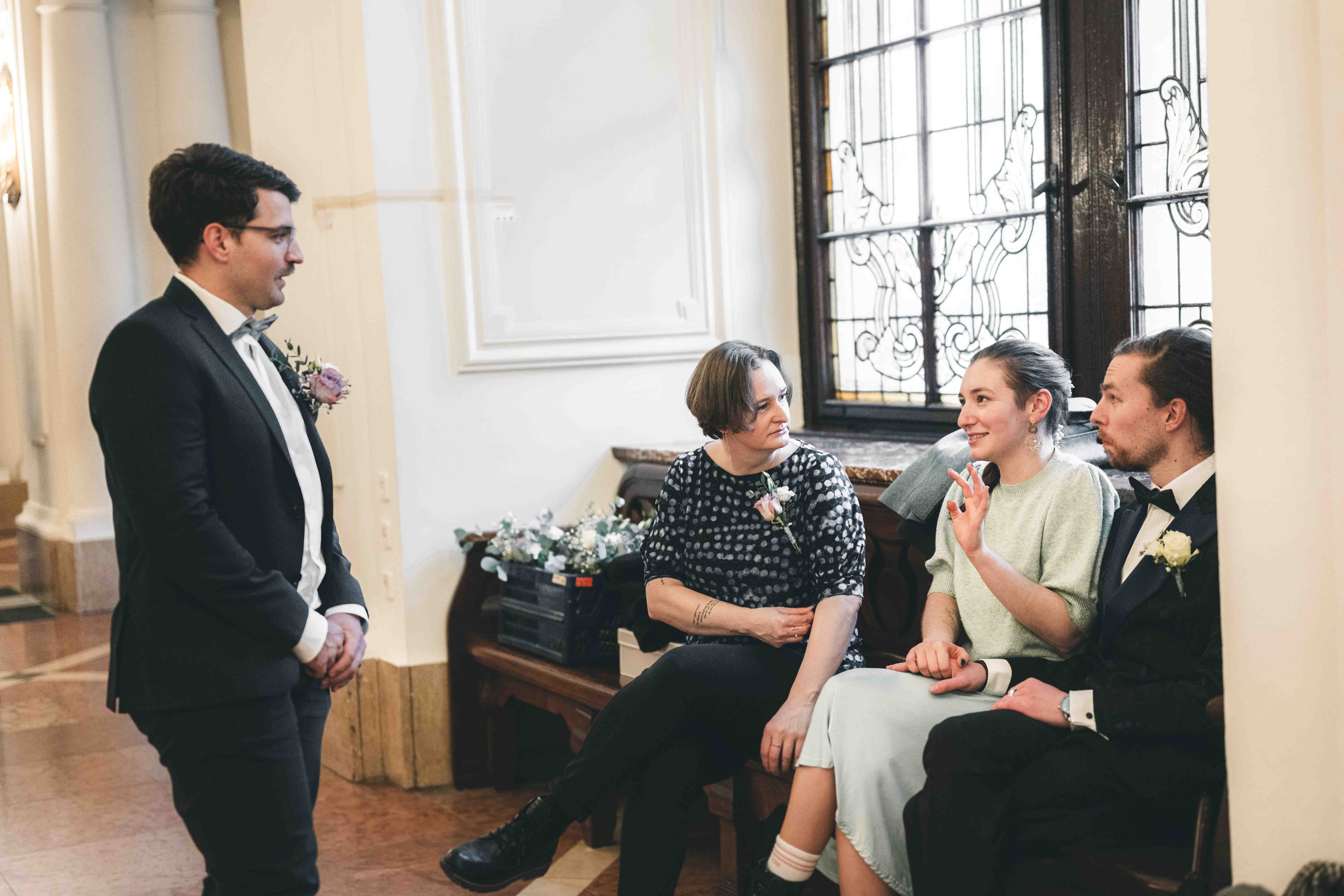 Eine Gruppe von Hochzeitsgästen unterhält sich entspannt auf einer Holzbank vor den historischen Buntglasfenstern im Neuen Rathaus Leipzig.