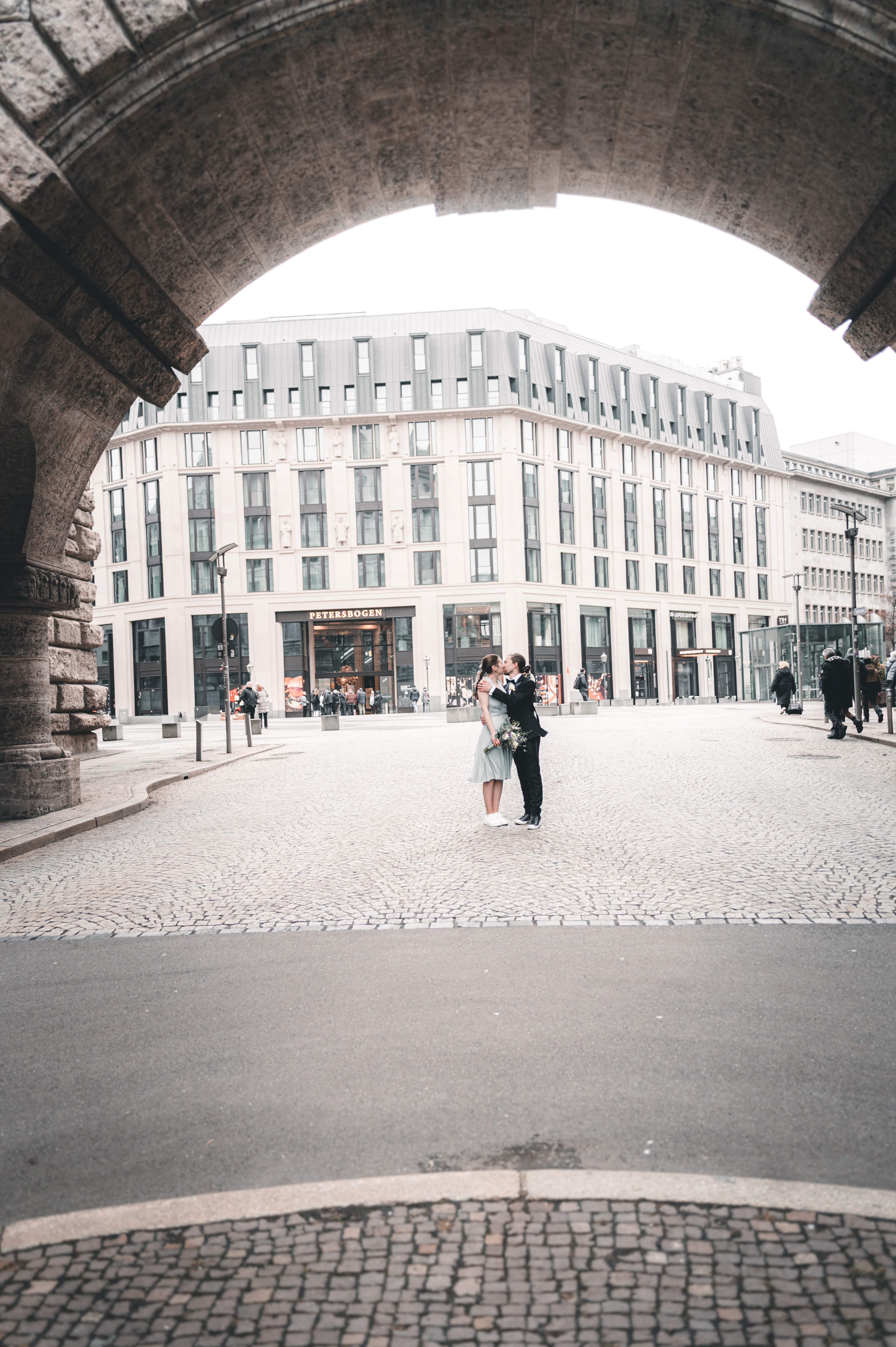 Ein weitwinkliger Blick durch einen gewaltigen Torbogen des Neuen Rathauses auf das Brautpaar, das sich auf dem gepflasterten Platz vor dem Petersbogen Leipzig küsst.