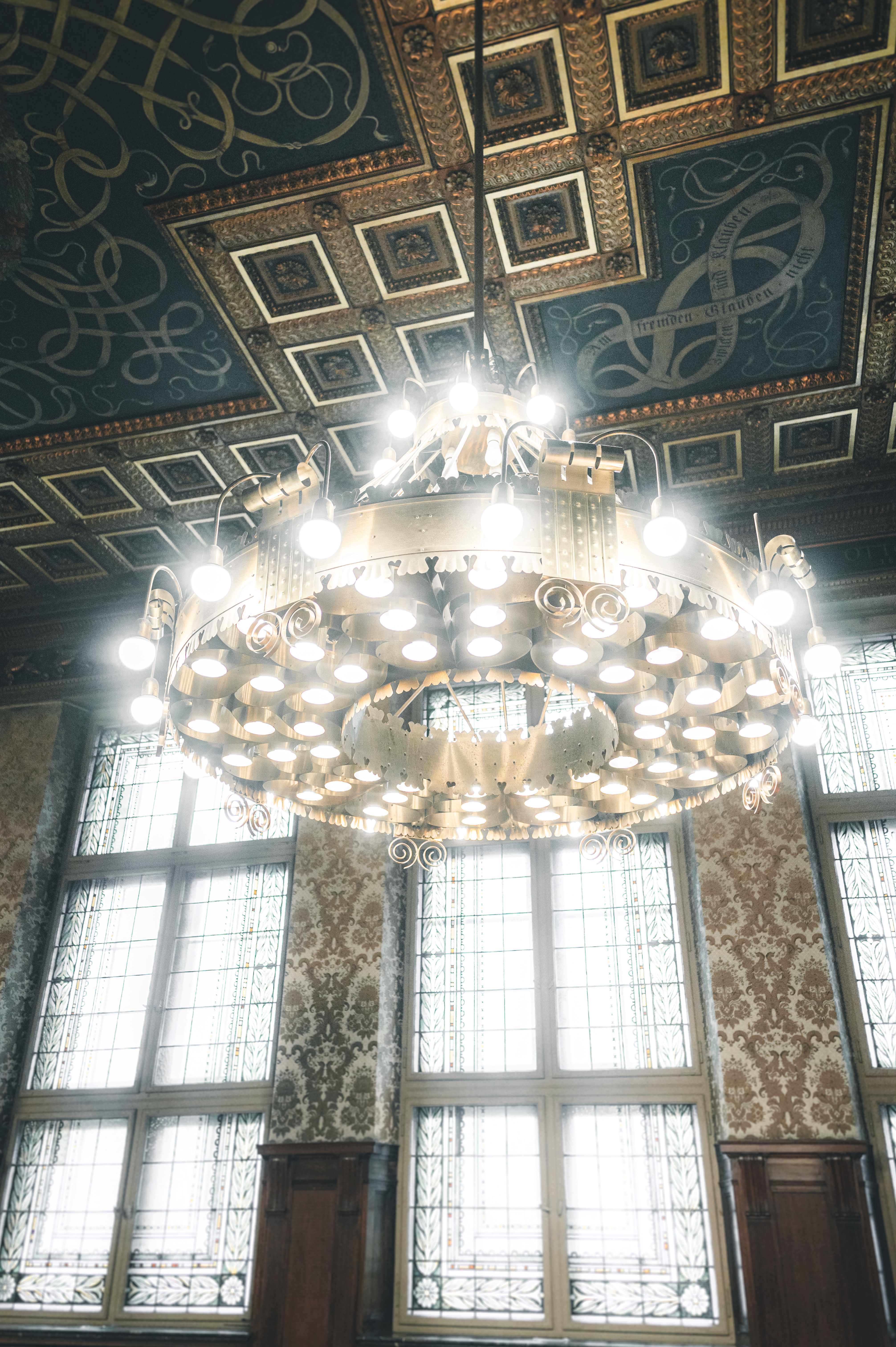 Ein riesiger, prunkvoller goldener Kronleuchter hängt unter der reich verzierten, dunklen Holzdecke des Ratsplenarsaals im Neuen Rathaus Leipzig.