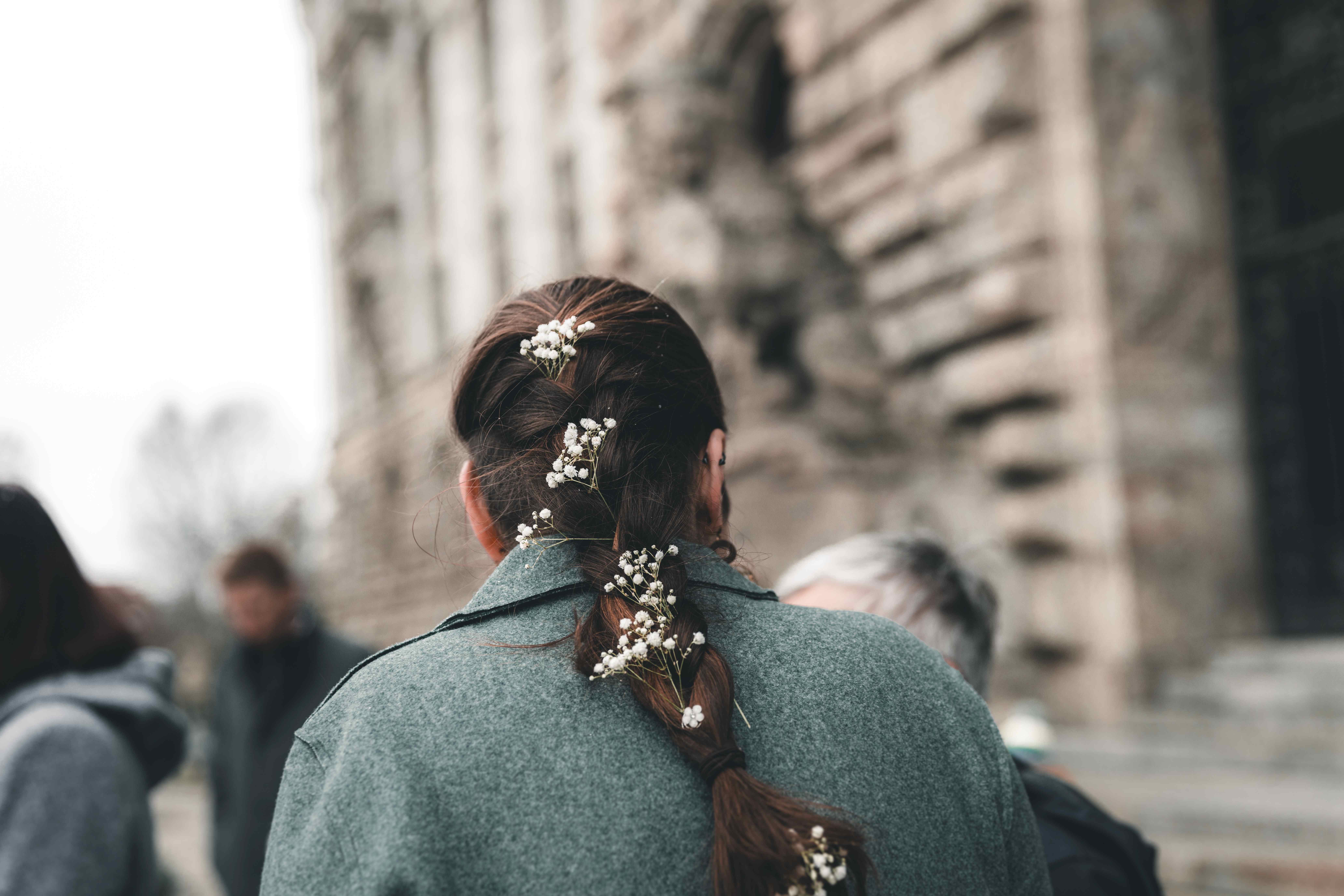 Die Braut im grünen Wintermantel von hinten vor der Steinfassade des Neuen Rathauses Leipzig, ihr geflochtener Zopf ist mit weißen Schleierkraut-Blüten verziert.