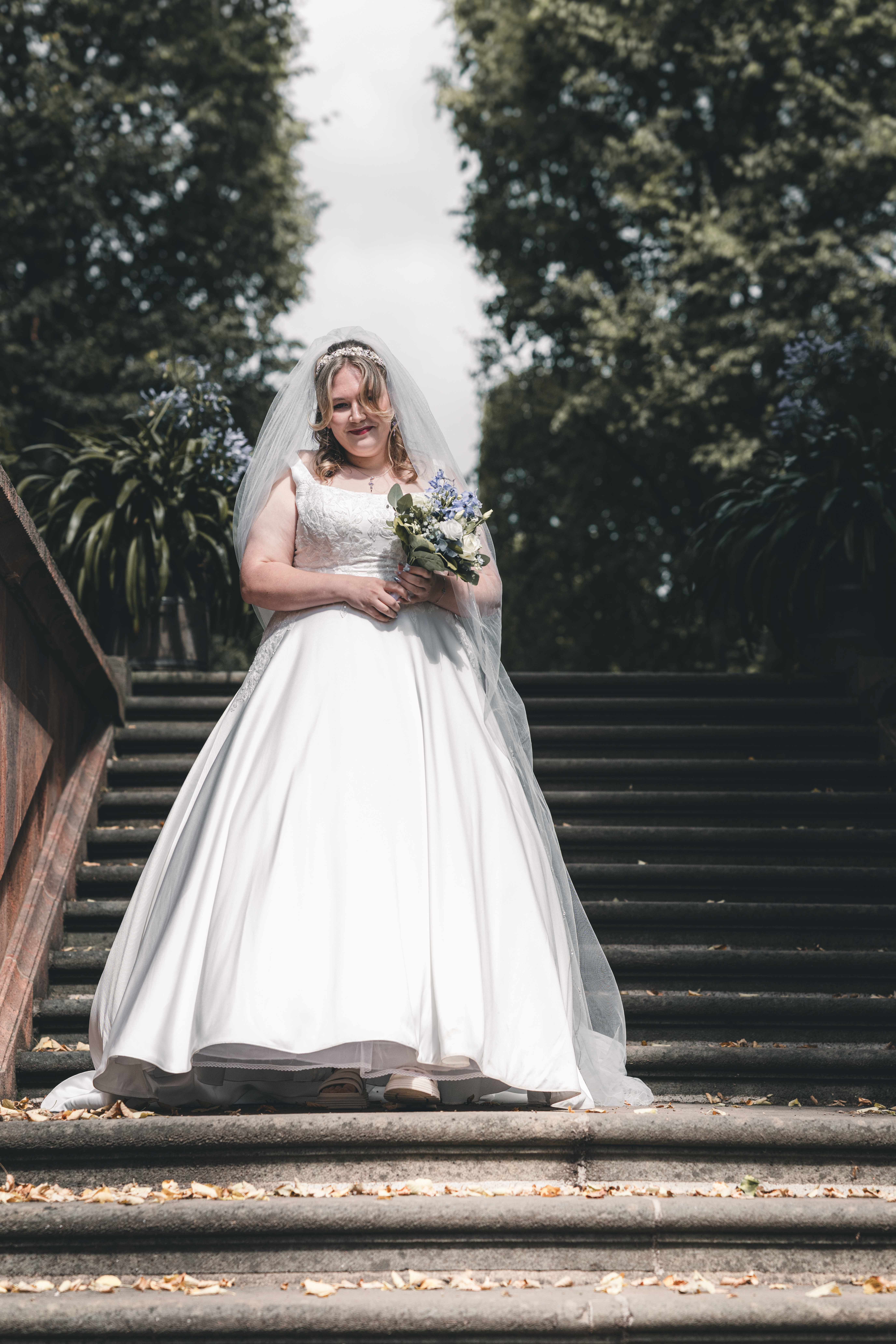 Die Braut im eleganten weißen Prinzessinnen-Brautkleid mit langem Schleier steht mit ihrem Strauß auf den Steinstufen im Schlosspark Lichtenwalde.