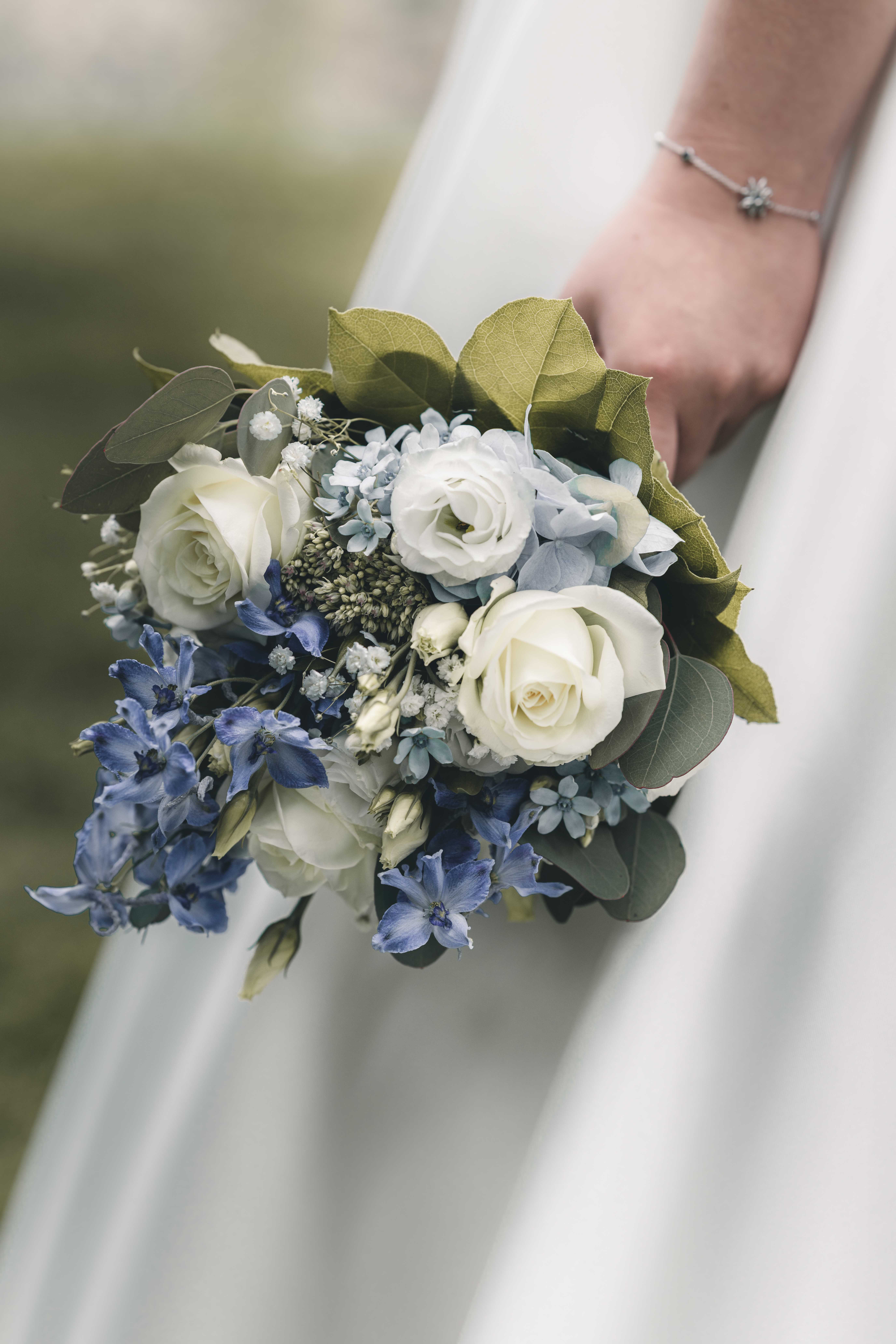 Nahaufnahme des Brautstraußes aus weißen Rosen, blauen Hortensien und Eukalyptus, gehalten von der Braut mit zartem Silberarmband im Raum Chemnitz.