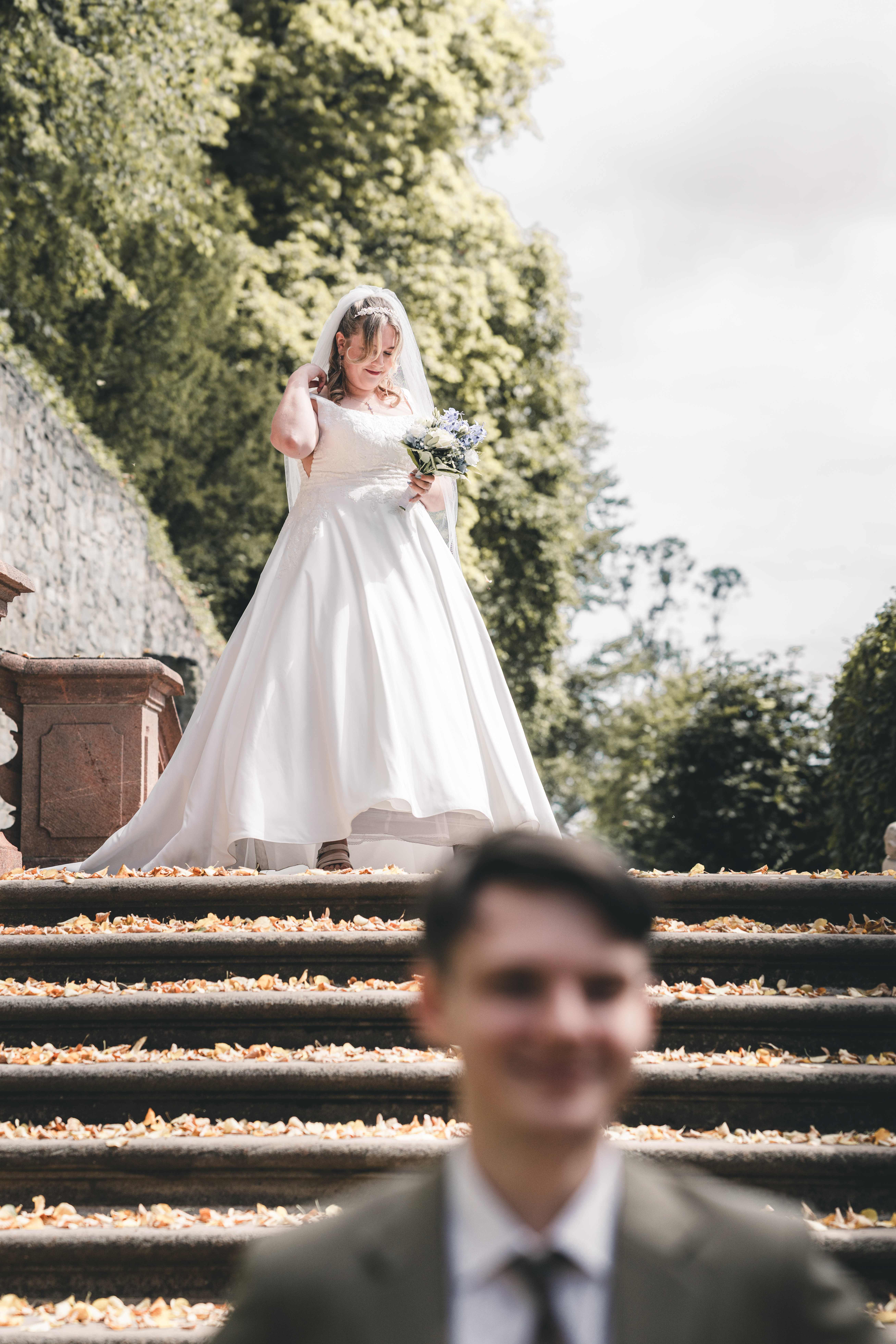 Die Braut im weißen Hochzeitskleid läuft mit ihrem Strauß die laubbedeckten Steinstufen im Schlosspark Lichtenwalde hinab, während der Bräutigam im Vordergrund unscharf wartet.