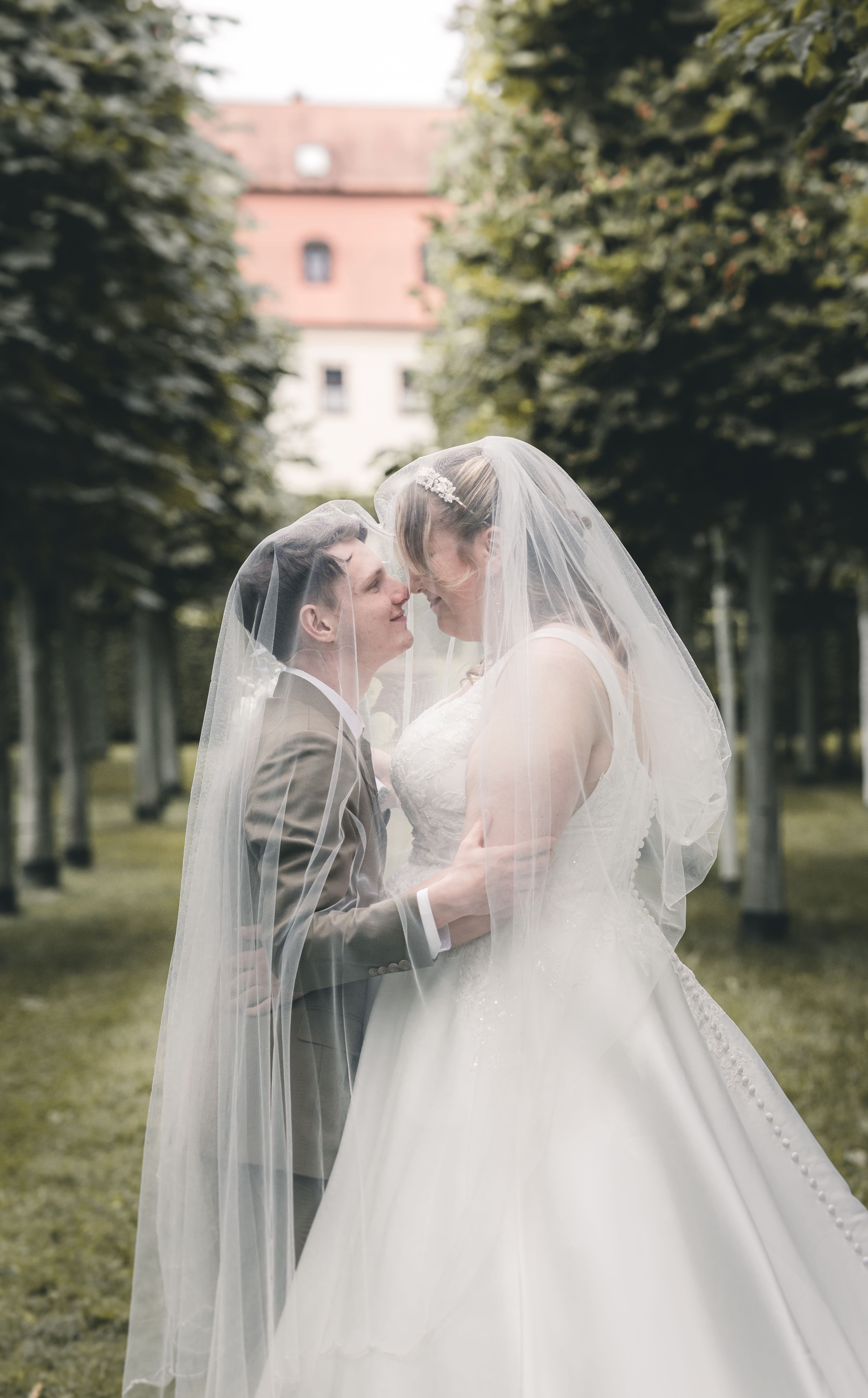 Eine romantische Detailaufnahme, bei welcher der Bräutigam seiner Braut unter dem fließenden Tüllschleier sanft einen Kuss auf die Stirn gibt.