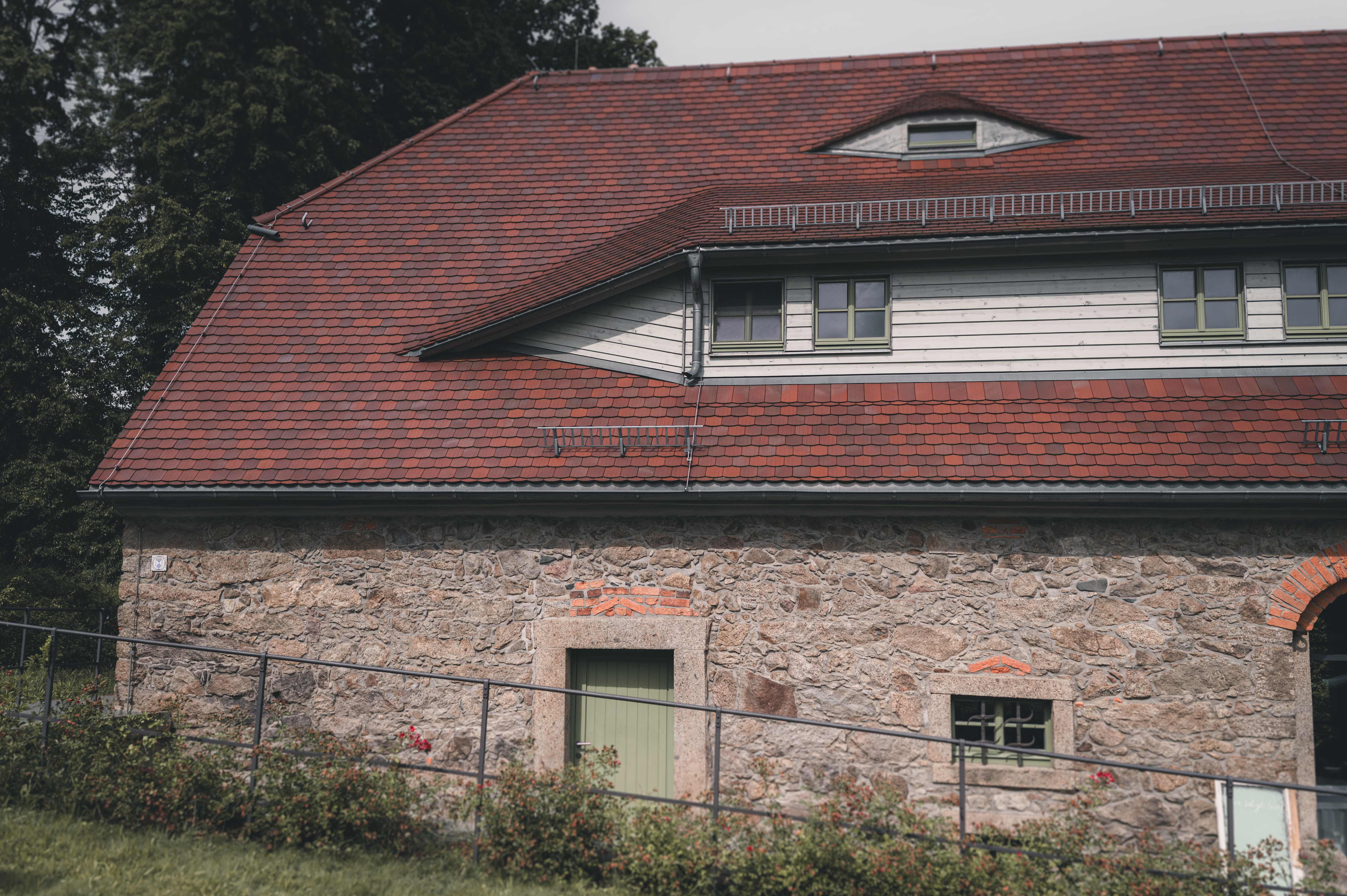 Die urige Natursteinfassade der Alten Schäferei in Oppach mit roten Dachziegeln und einer von Rosen gesäumten Holzgalerie.
