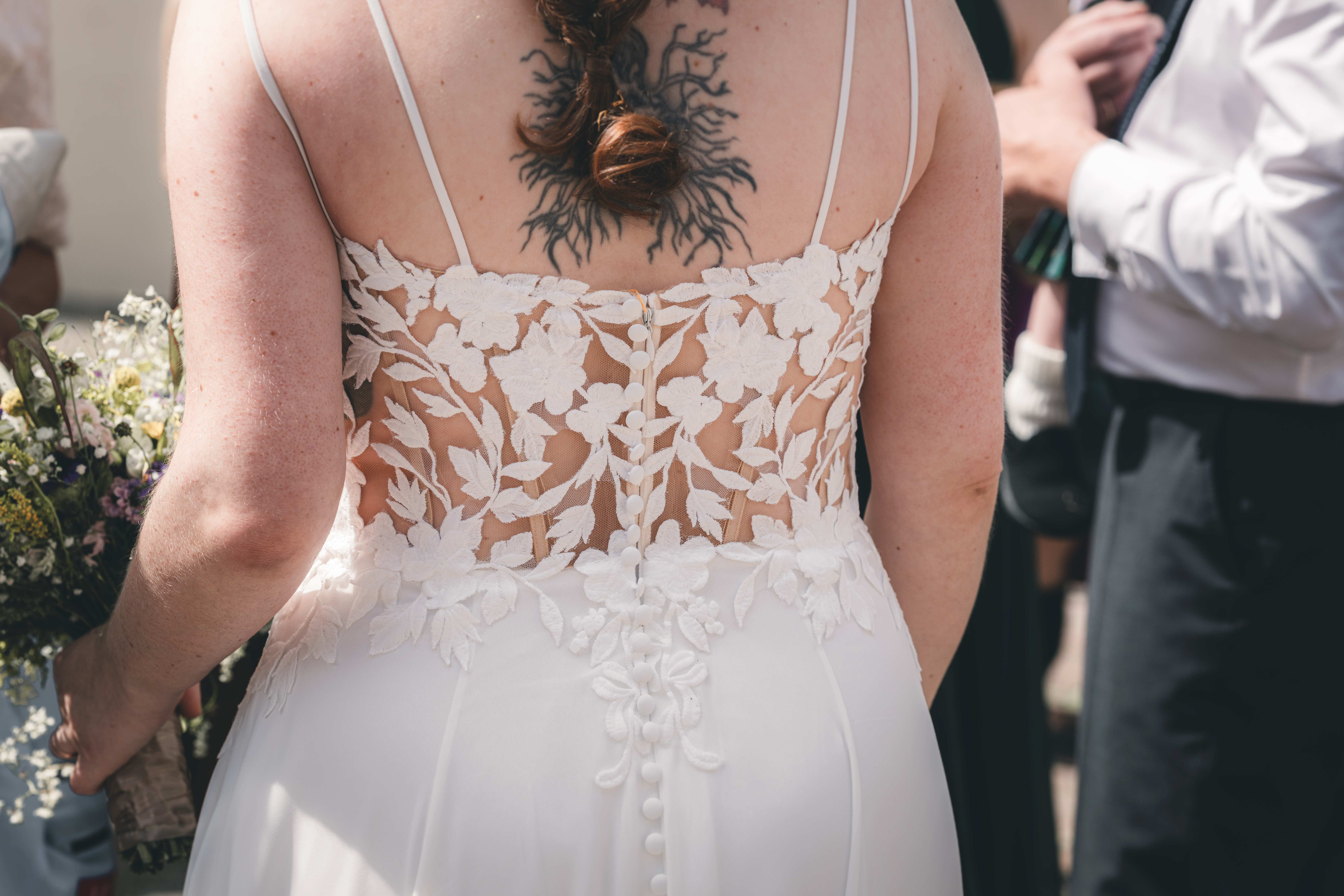 Nahaufnahme der filigranen floralen Spitze am Rücken des Hochzeitskleides der Braut, die ein kunstvolles Tattoo im Nacken zeigt.