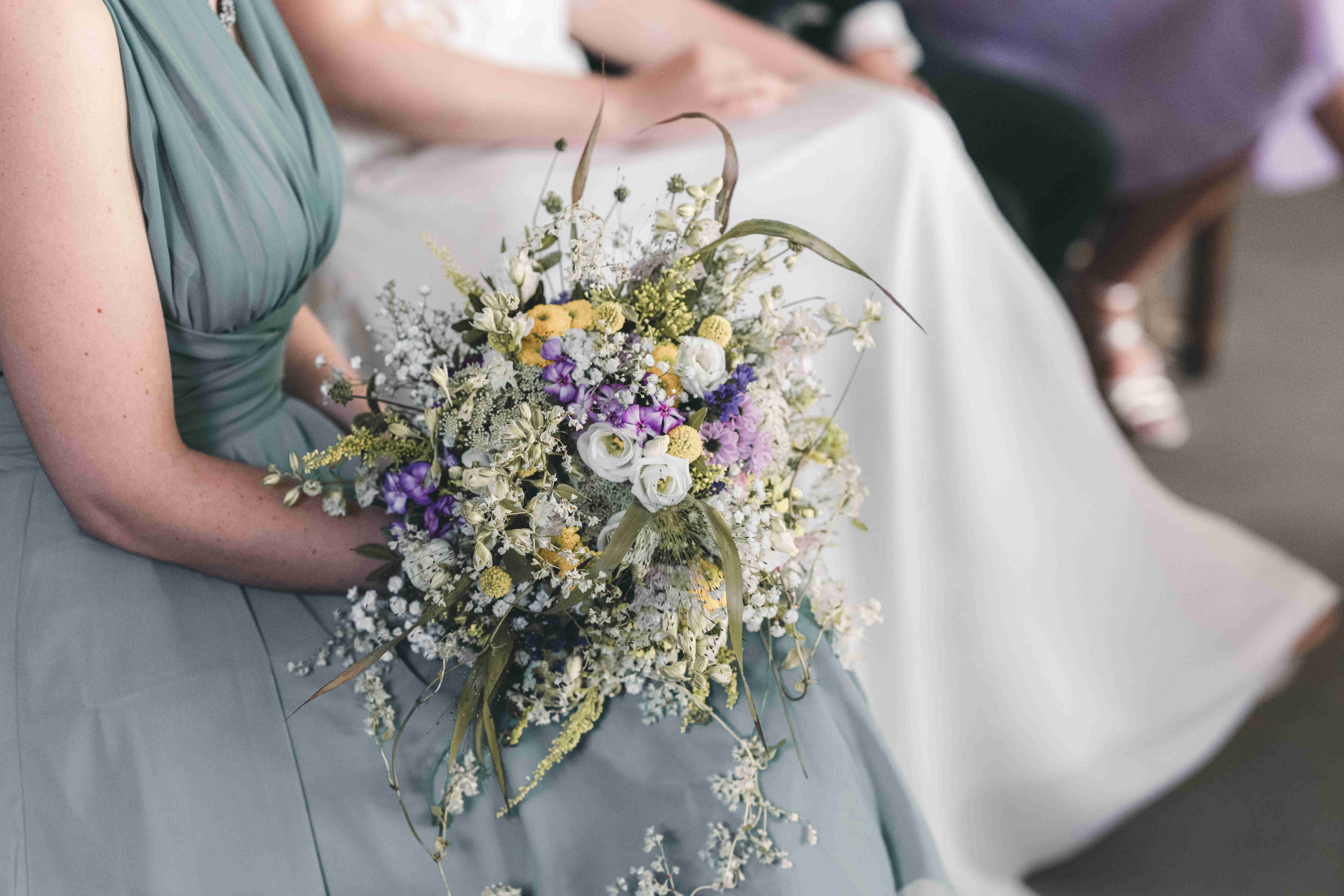 Eine Nahaufnahme des sommerlichen Brautstraußes aus Wiesenblumen, der zart auf dem Kleid einer Brautjungfer liegt.