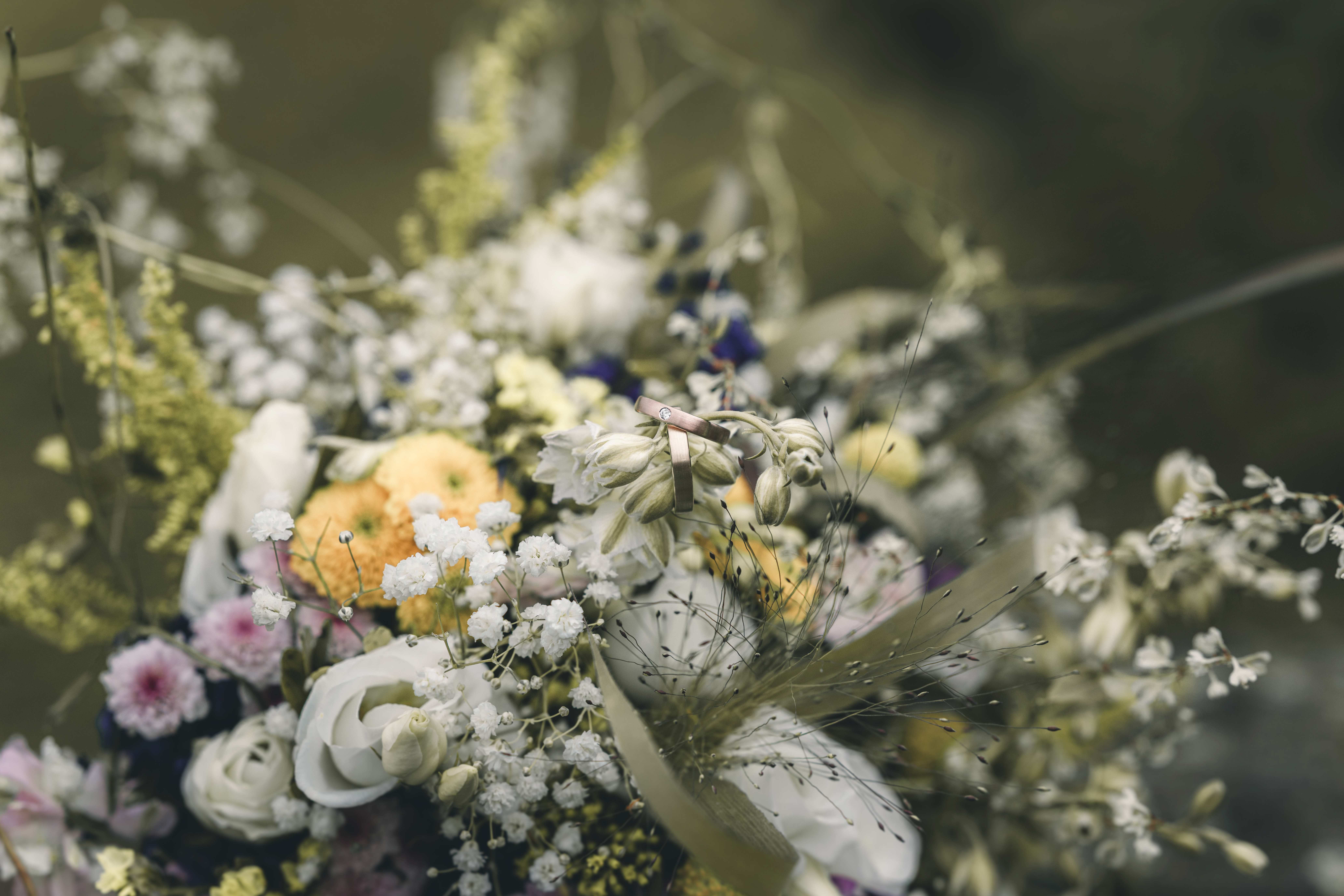 Eine detailreiche Makroaufnahme der beiden Eheringe, die filigran an den Knospen des Sommerstraußes aus Schleierkraut und Wiesenblumen hängen.
