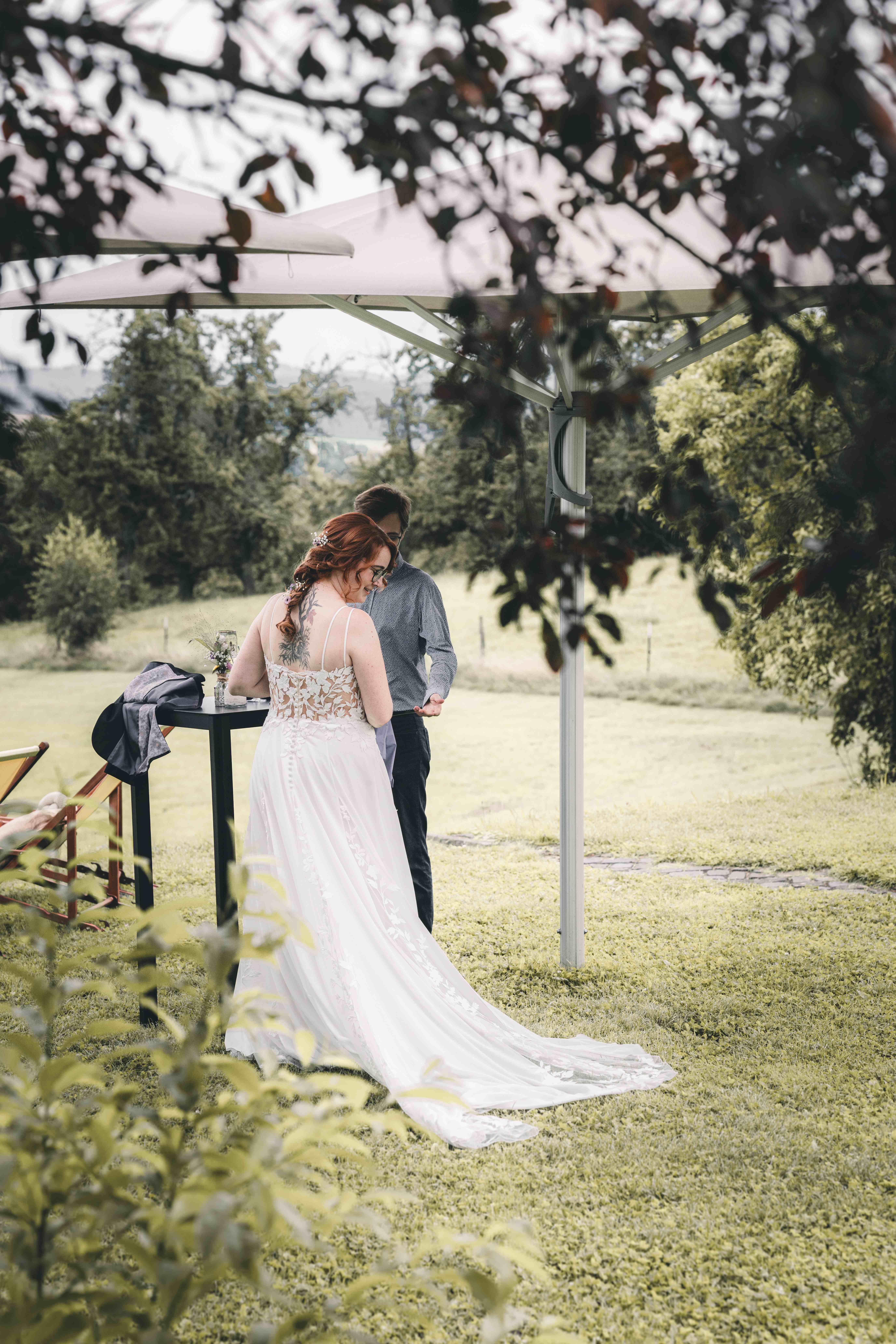 Die Braut im eleganten Spitzenkleid mit langer Schleppe steht unter einem Sonnenschirm auf der Festwiese der Alten Schäferei in Oppach.