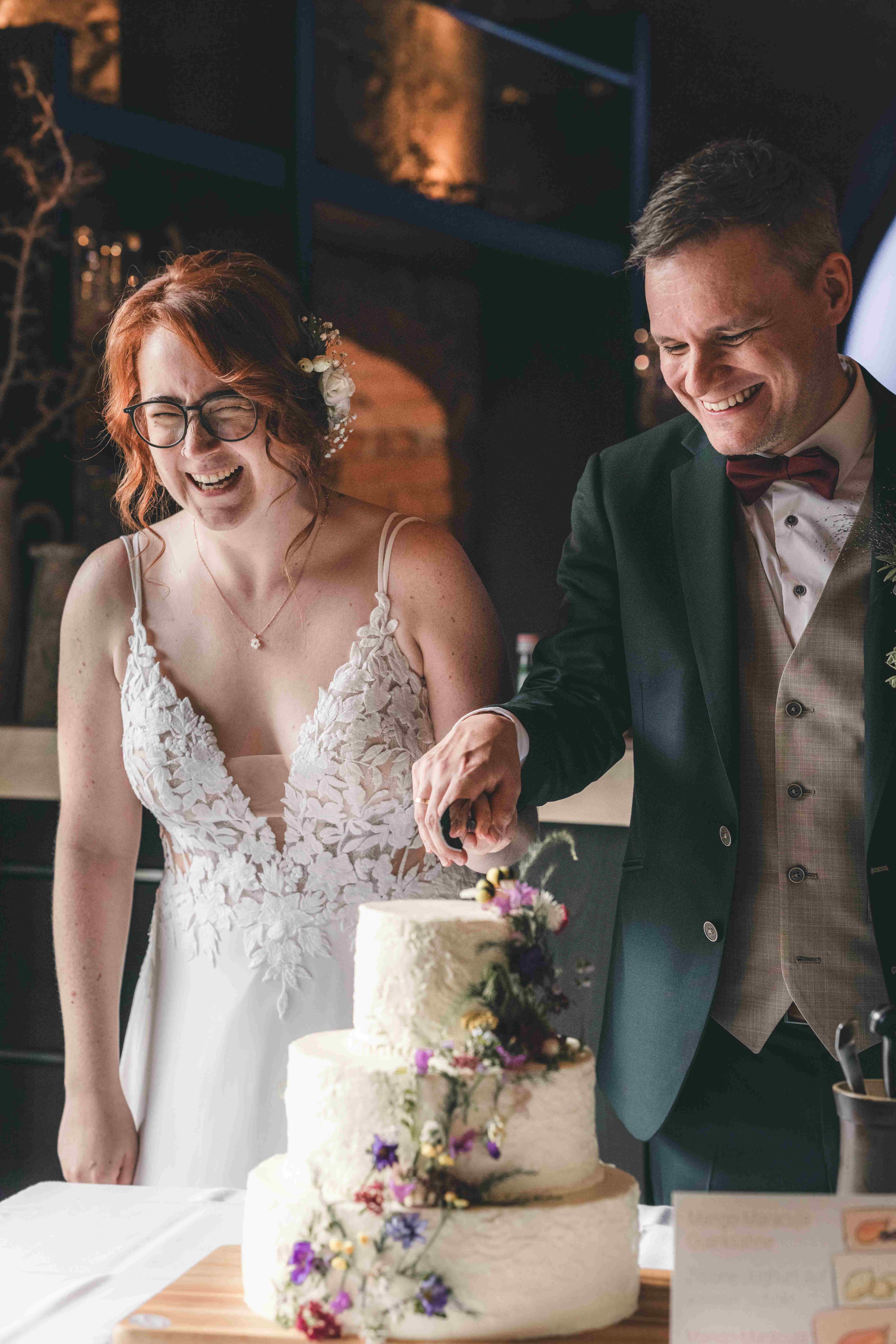 Die Braut lacht herzlich, während das Paar gemeinsam das Messer durch die Hochzeitstorte führt – ein lebendiger Moment der Hochzeitsfeier.