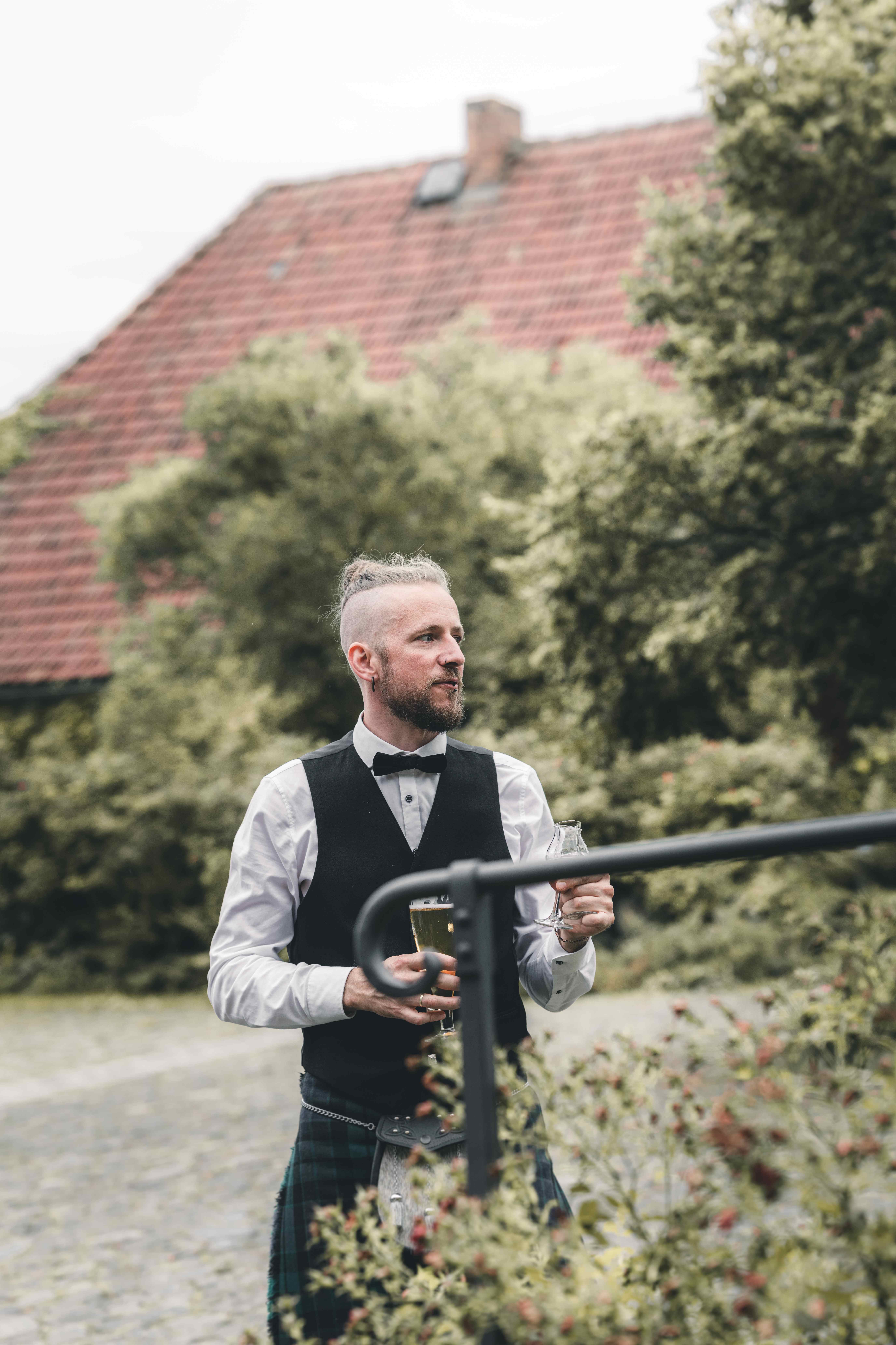 Ein Hochzeitsgast in einem traditionellen schottischen Kilt und einer schwarzen Weste hält ein Glas in der Hand und schaut lächelnd in den Garten.