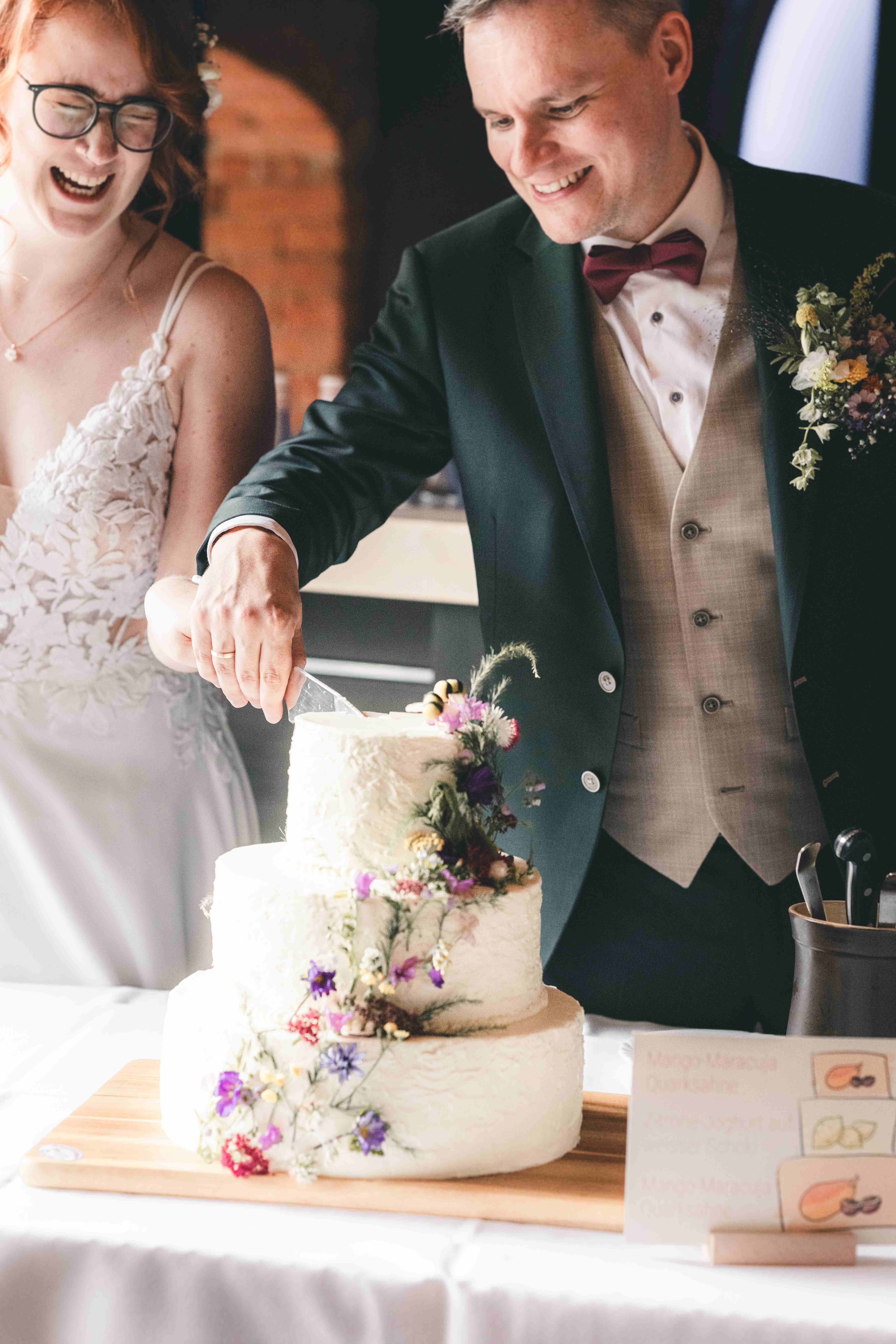 Das Brautpaar schneidet gemeinsam lächelnd die dreistöckige Hochzeitstorte mit Wiesenblumen-Deko in der Alten Schäferei Oppach an.