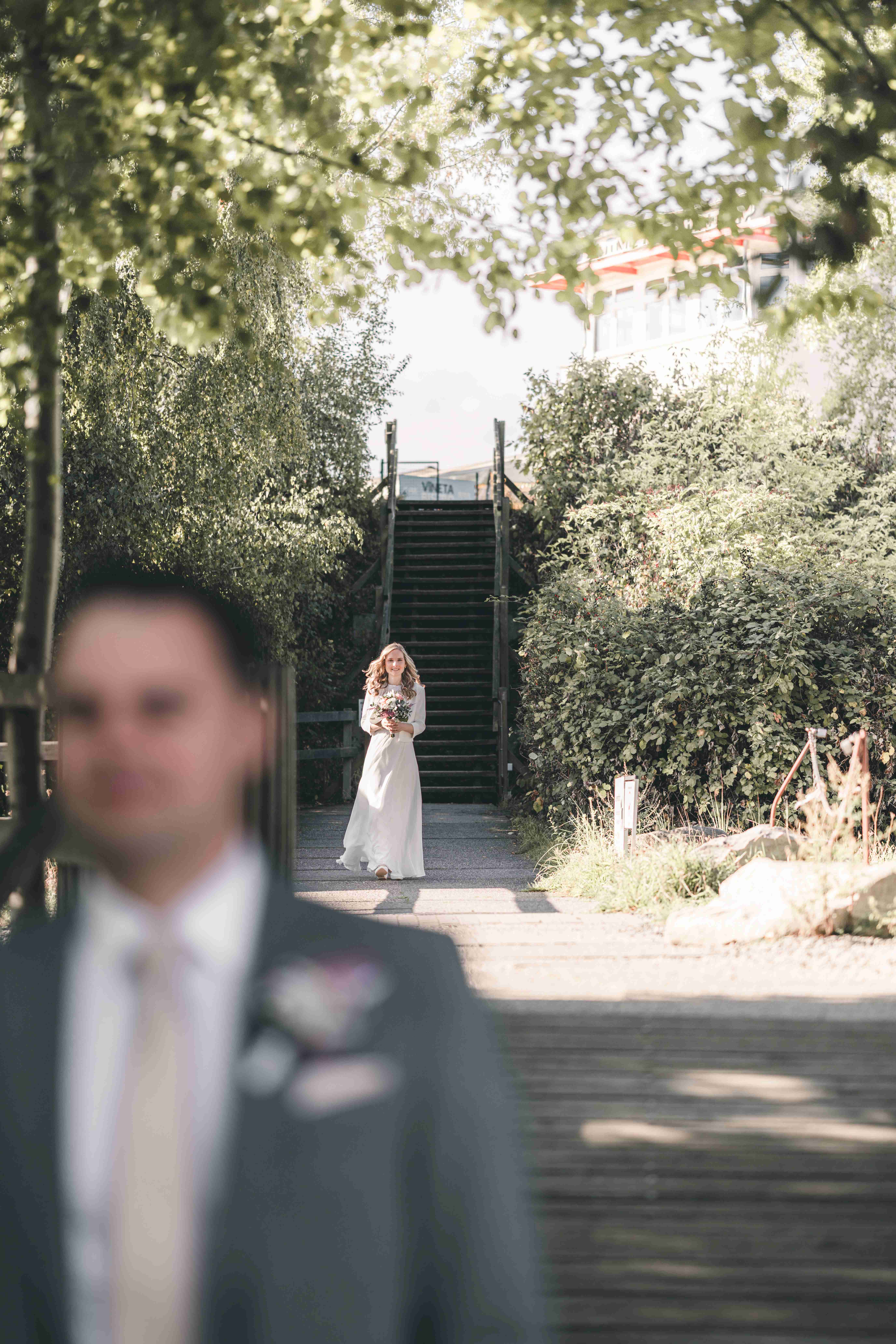 Die Braut im weißen Hochzeitskleid läuft mit ihrem Strauß eine hölzerne Treppe hinab, während der Bräutigam im Vordergrund unscharf wartet.