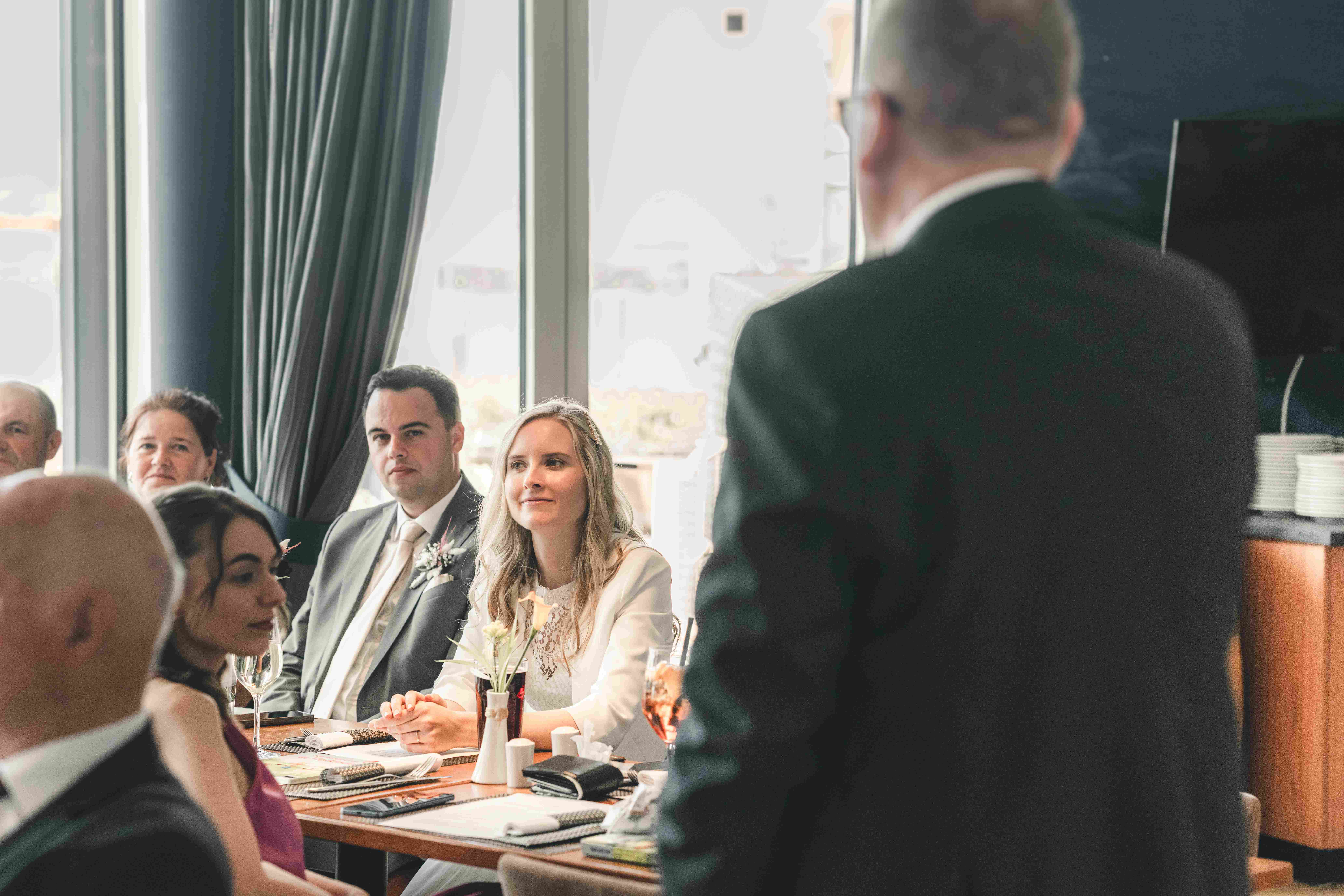 Das Brautpaar sitzt am Hochzeitstisch im Hotel Lagovida und blickt gerührt zum Redner, während der Brautvater im Vordergrund unscharf zu sehen ist.